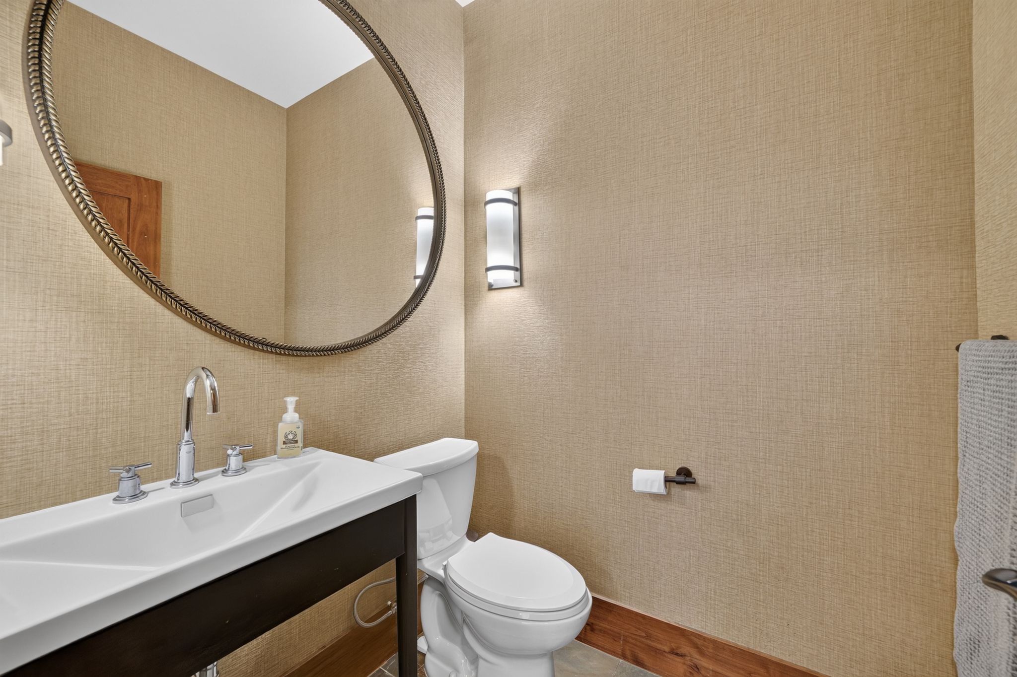 Powder room featuring a sleek white rectangular sink, chrome fixtures, a large oval mirror with an ornate frame, textured wall covering, and two sconce light fixtures