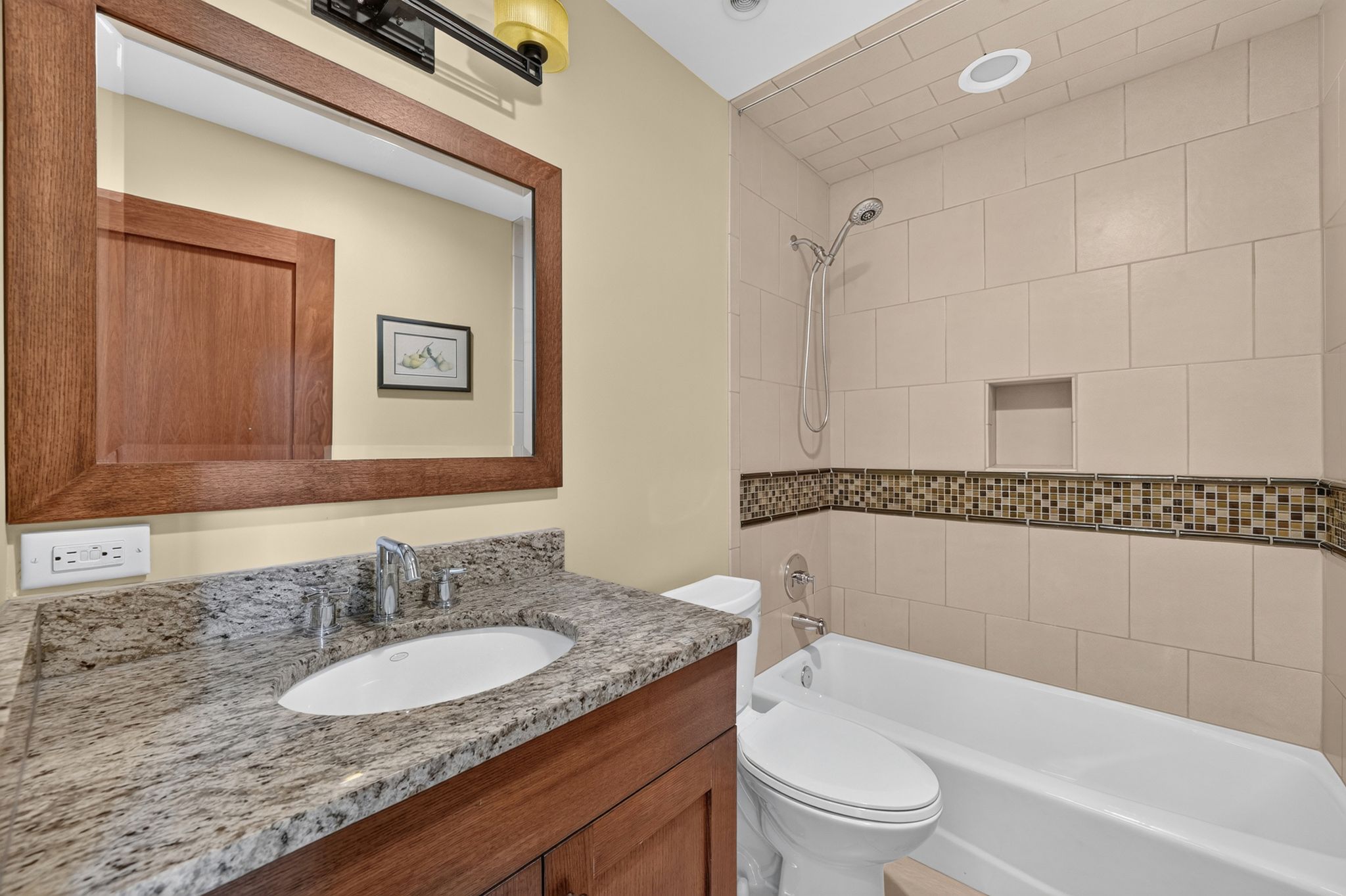 Bathroom featuring a wood-framed mirror, integrated sink with a stone countertop, wood-finish vanity cabinetry, and a full-size bathtub with subway-style tiling and a mosaic accent band