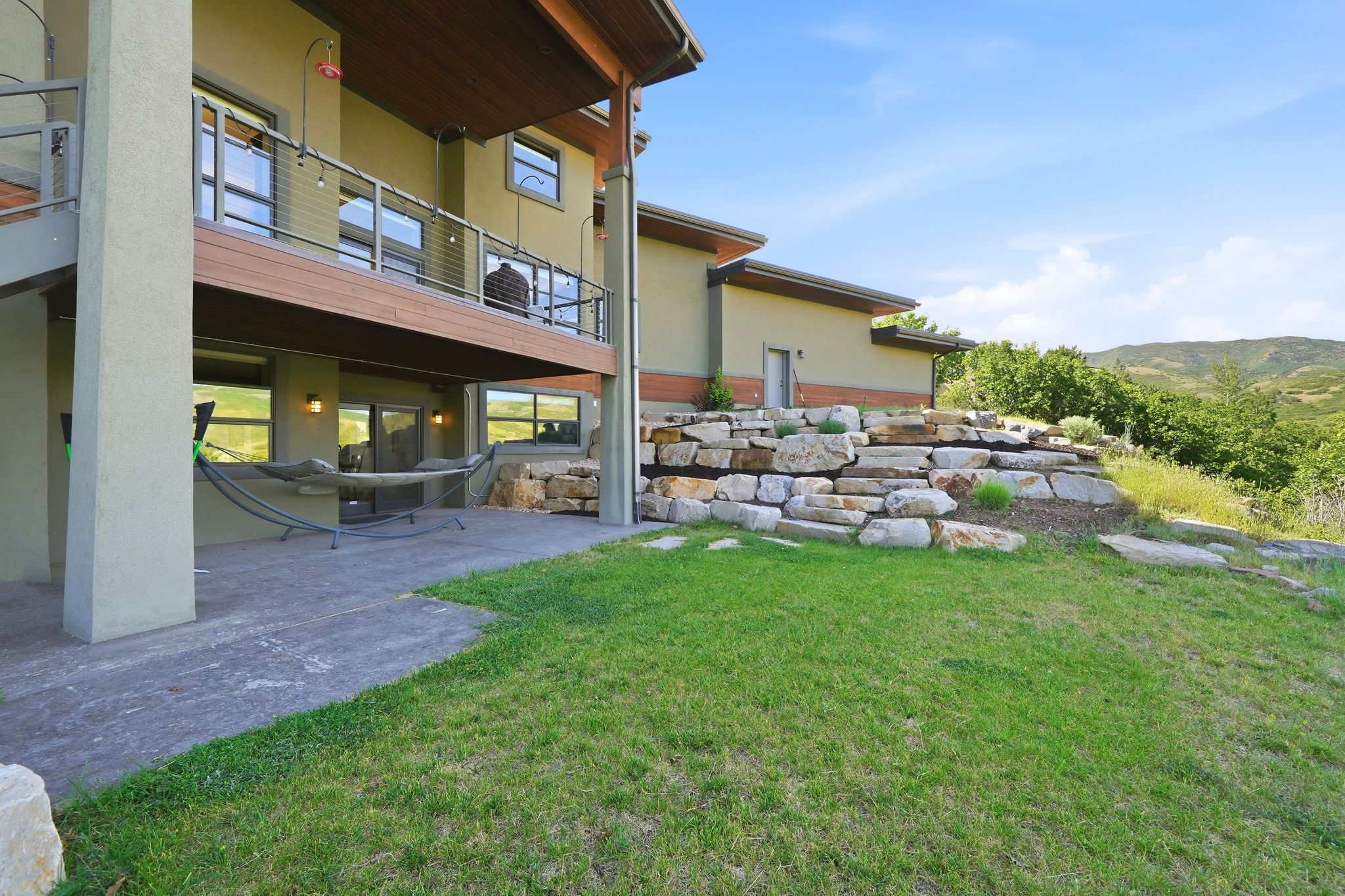 Rear exterior featuring a large concrete patio, an upper-level balcony with cable railings, stucco siding, and extensive stone terracing
