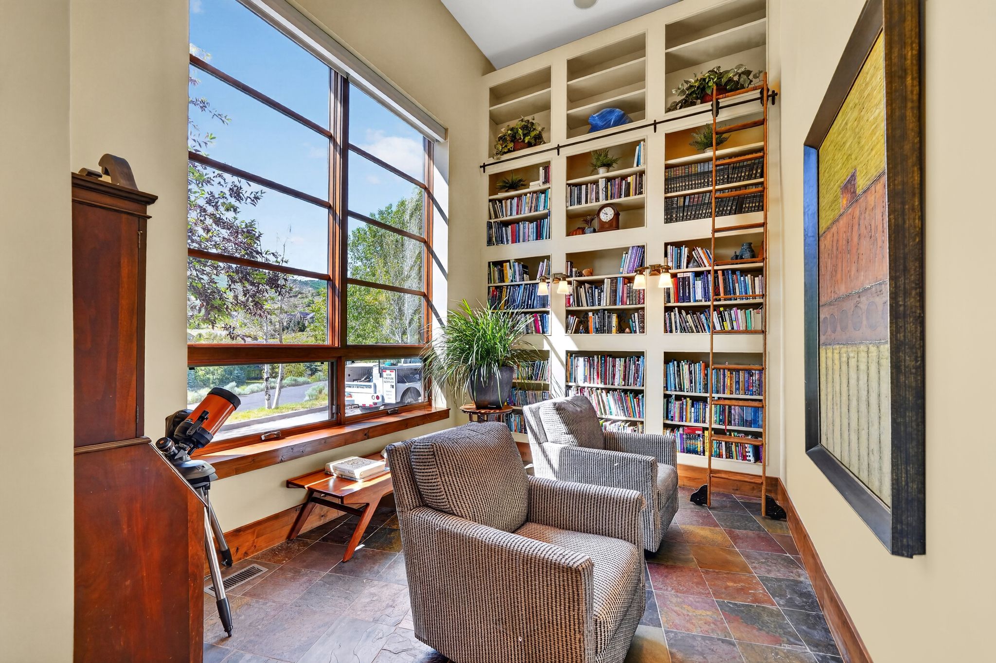 Spacious room featuring a multi-pane picture window, floor-to-ceiling built-in shelving with a rolling ladder, and multi-tone tile flooring