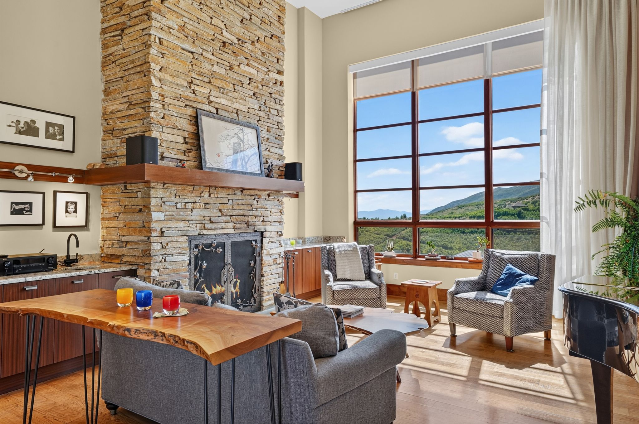 Two-story living area featuring a stacked stone fireplace with a wood mantle, a wet bar with a dark faucet, and a large multi-pane picture window