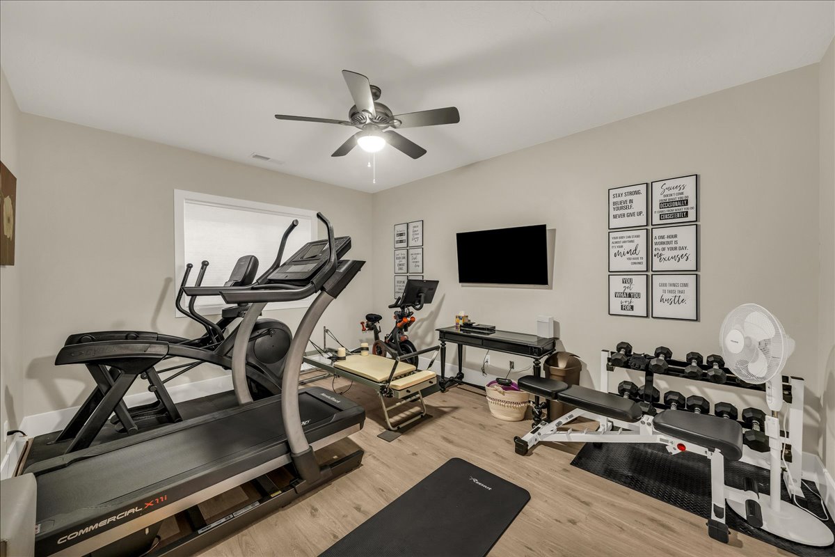 Dedicated fitness space featuring wood-finish flooring, neutral wall tones, a ceiling fan with integrated lighting, and a white-trimmed window