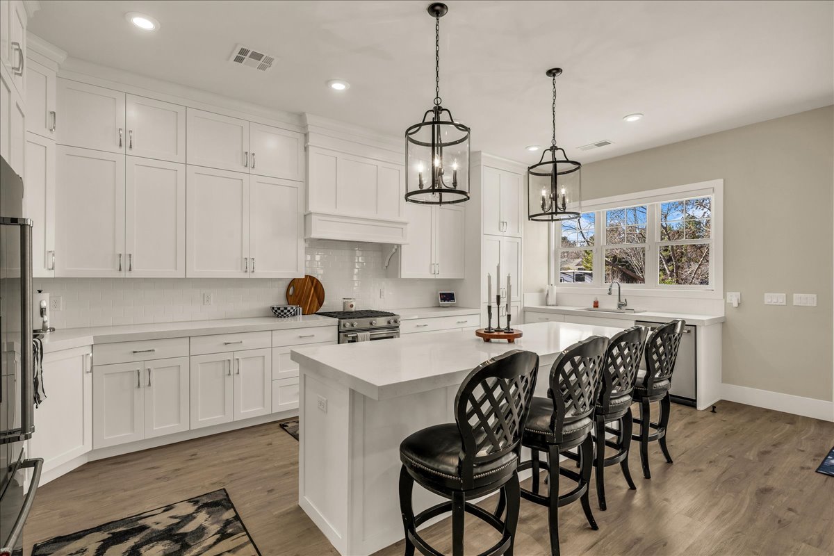 Bright kitchen featuring white shaker cabinetry, an expansive central island with seating, stainless steel appliances, wood-finish flooring, and two modern pendant light fixtures