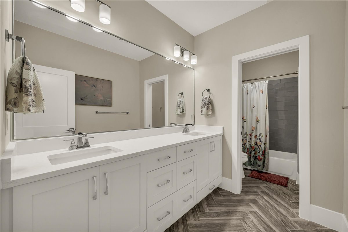 Bathroom featuring a spacious double vanity with white cabinetry and integrated sinks, a large wall-mounted mirror, overhead bar lighting, and wood-finish flooring laid in a herringbone pattern