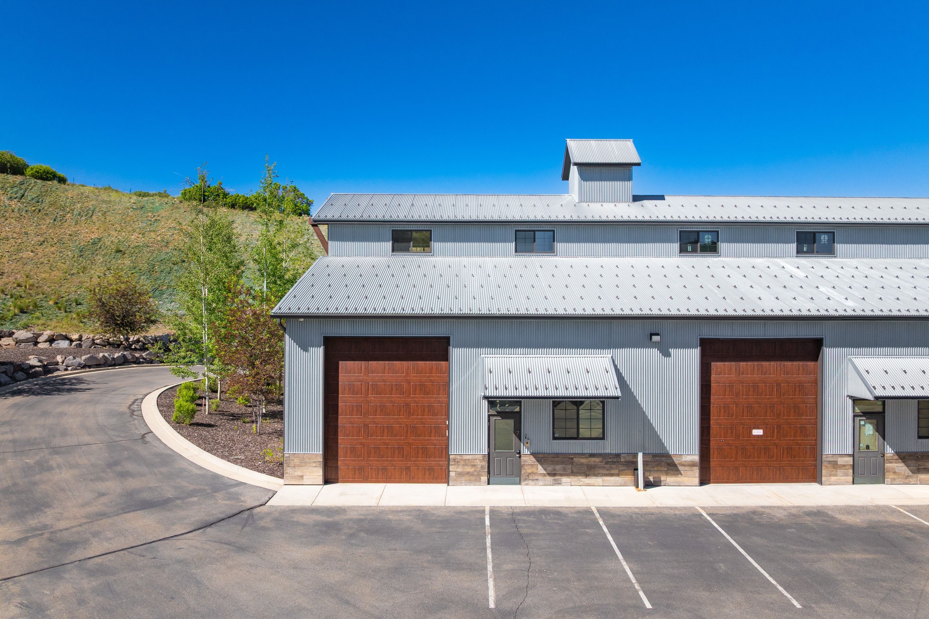 Modern industrial-style structure featuring a light gray exterior, two expansive brown garage doors, multiple windows with awnings, a cupola, and a paved parking area