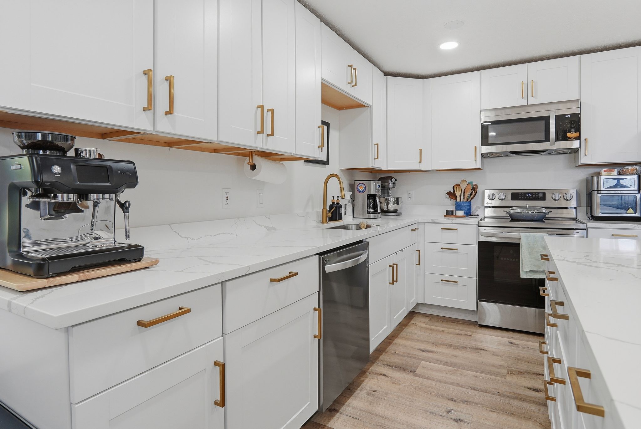 Modern kitchen featuring white shaker cabinetry, stainless steel appliances, quartz countertops, wood-finish flooring, and a gold-tone faucet