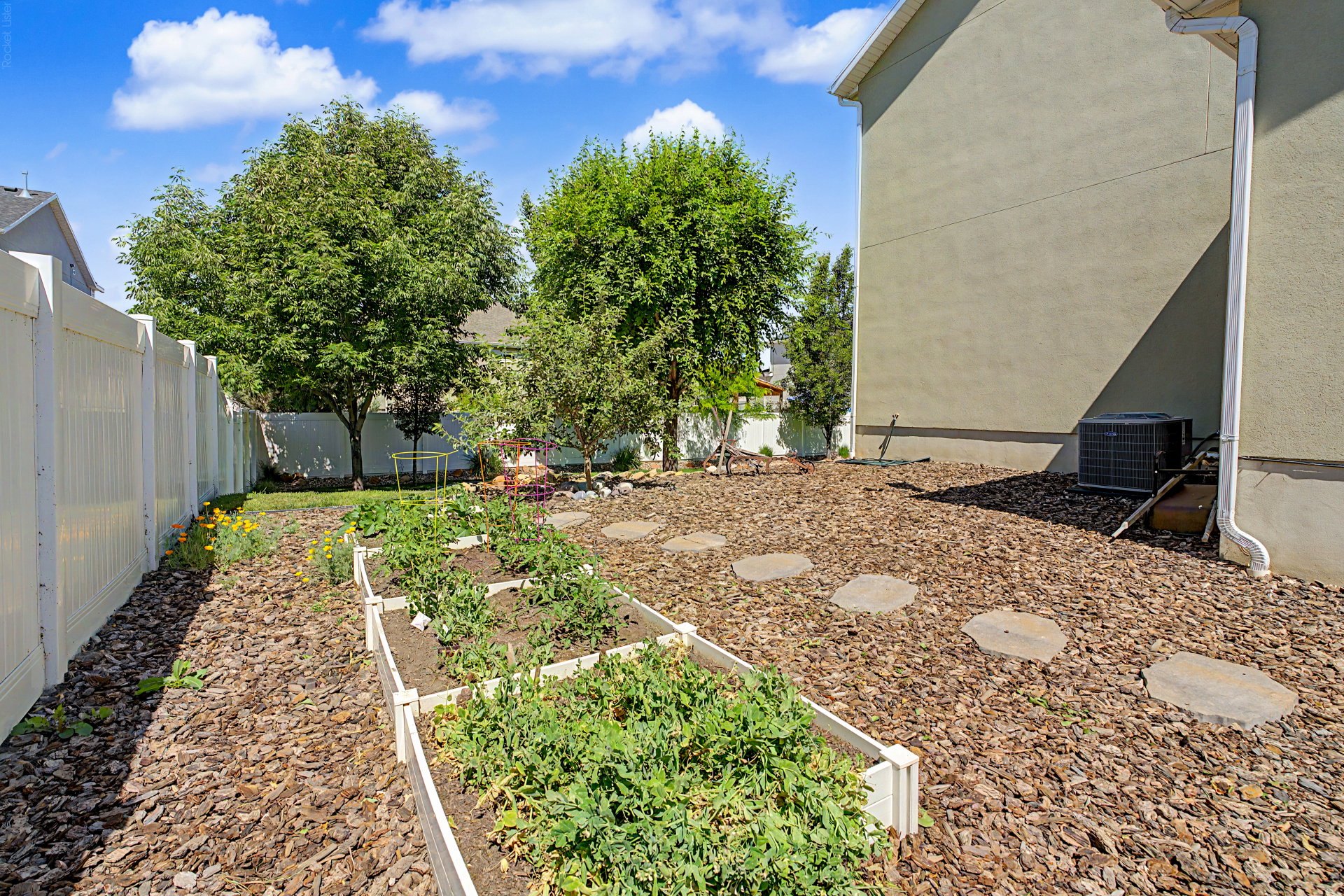Expansive backyard featuring multiple raised garden beds, a white vinyl privacy fence, and mature trees