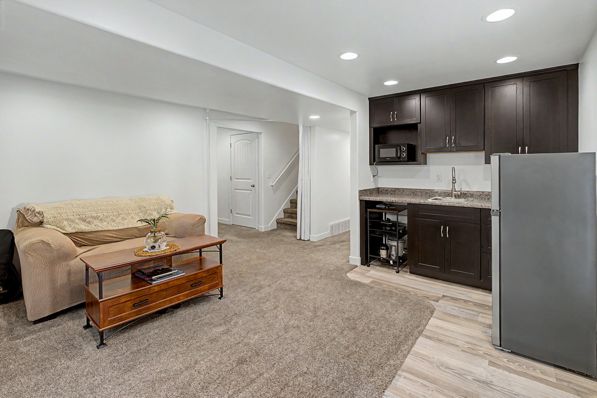 Lower level living area with a built-in kitchenette featuring dark wood cabinetry, a stainless steel sink, and a stone-finish countertop