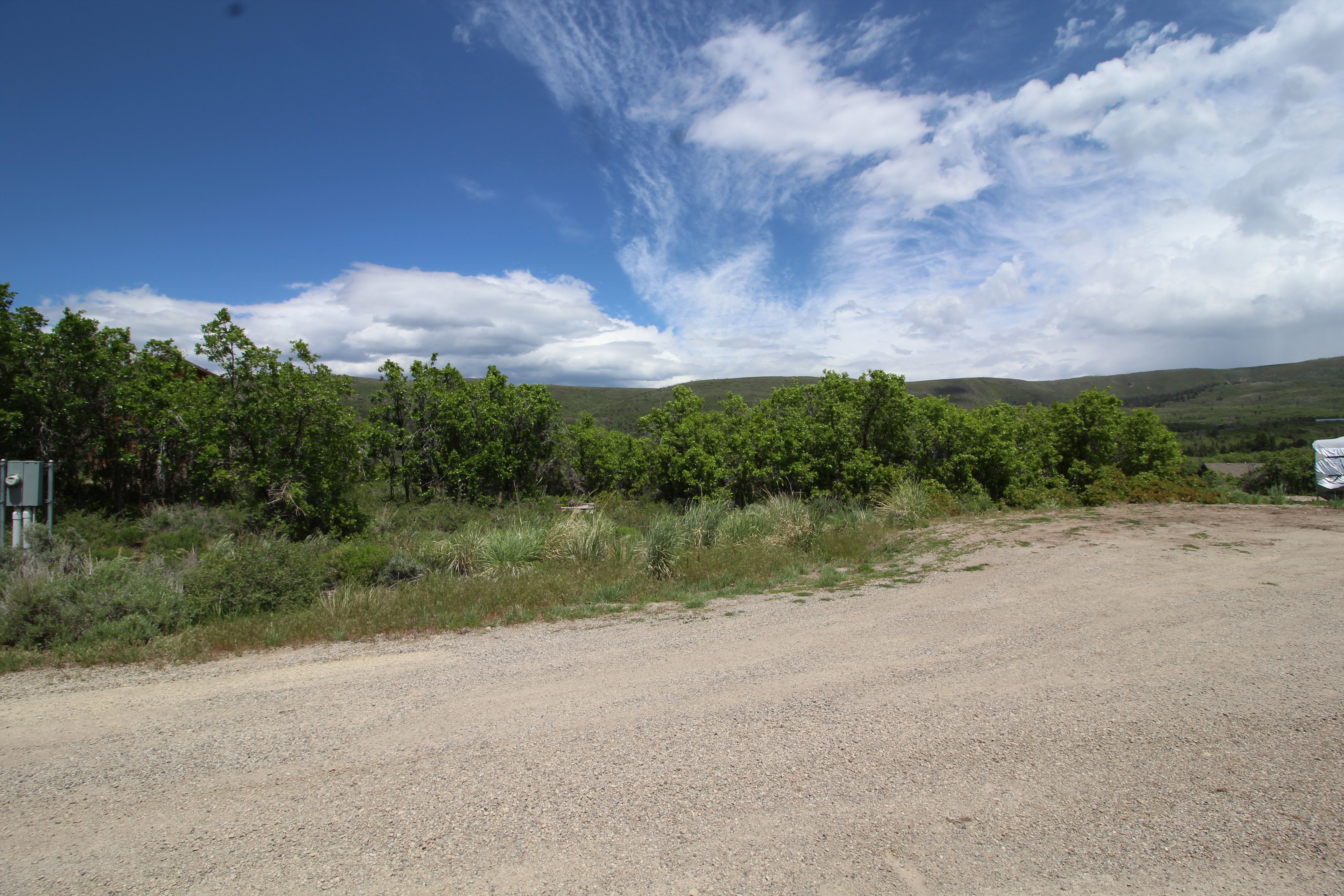 Expansive undeveloped lot featuring a gravel surface, natural vegetation, and distant rolling hills