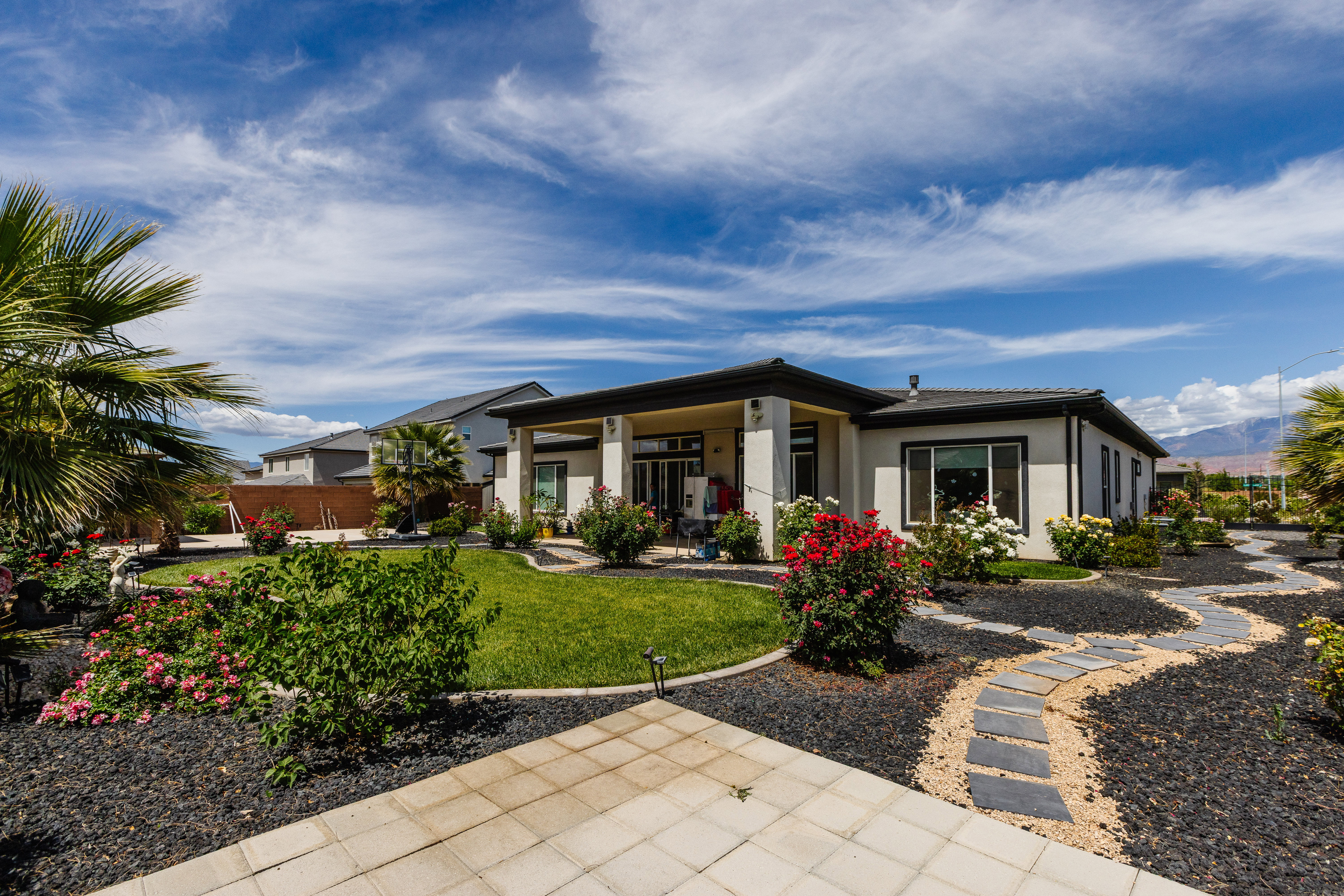 Spacious backyard featuring a covered patio, manicured lawn, paver stone walkway, mature landscaping, and decorative rock beds