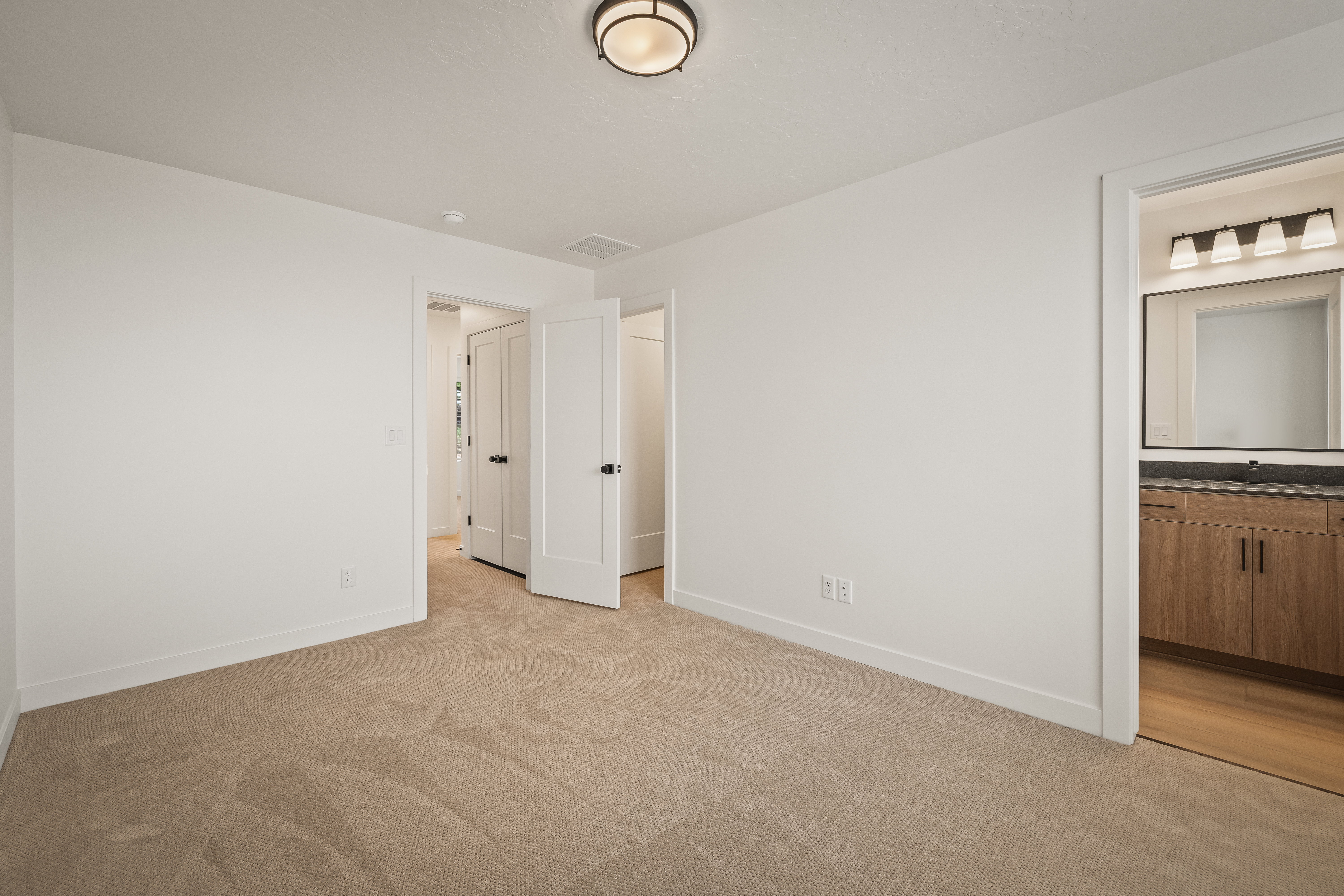 Carpeted room featuring a ceiling-mounted light fixture, white trim, and a doorway to a bathroom with a wood-finish vanity, dark countertop, and rectangular mirror with a four-light fixture