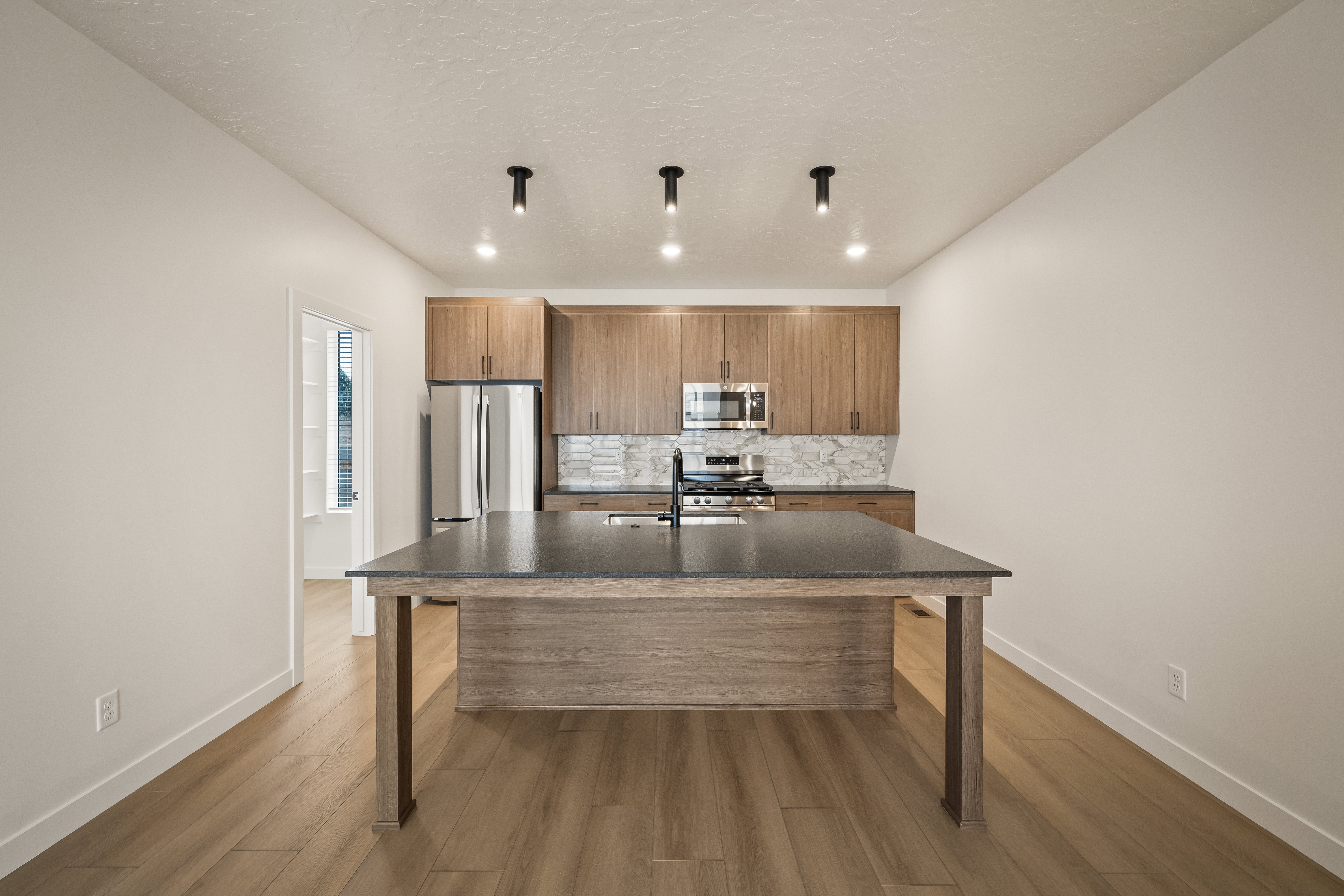Modern kitchen featuring an expansive center island with a dark countertop, stainless steel appliances, wood-finish cabinetry, a patterned tile backsplash, and light wood-finish flooring
