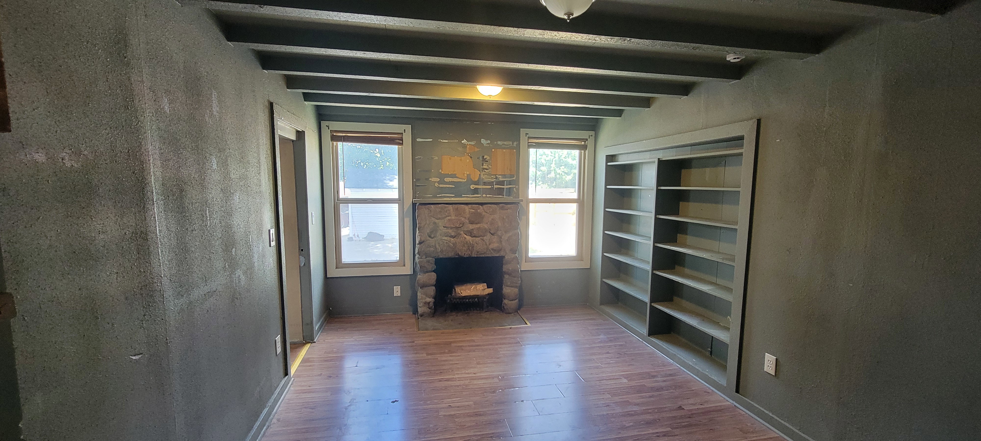 Stone fireplace flanked by two windows, built-in shelving, exposed ceiling beams, and wood-finish flooring
