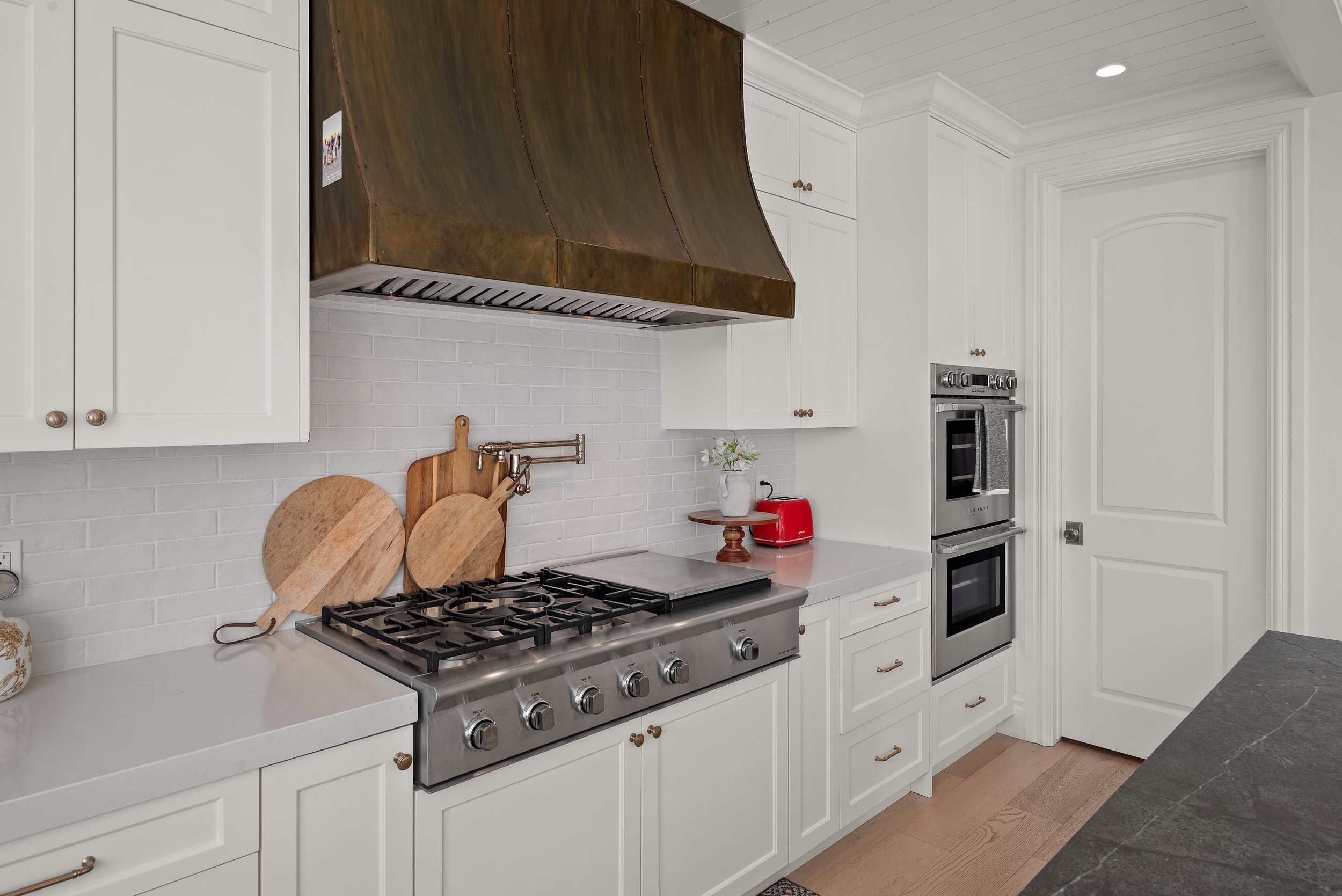 Gourmet kitchen featuring a substantial brass range hood, stainless steel gas cooktop, double wall ovens, white shaker cabinetry, and light-toned countertops