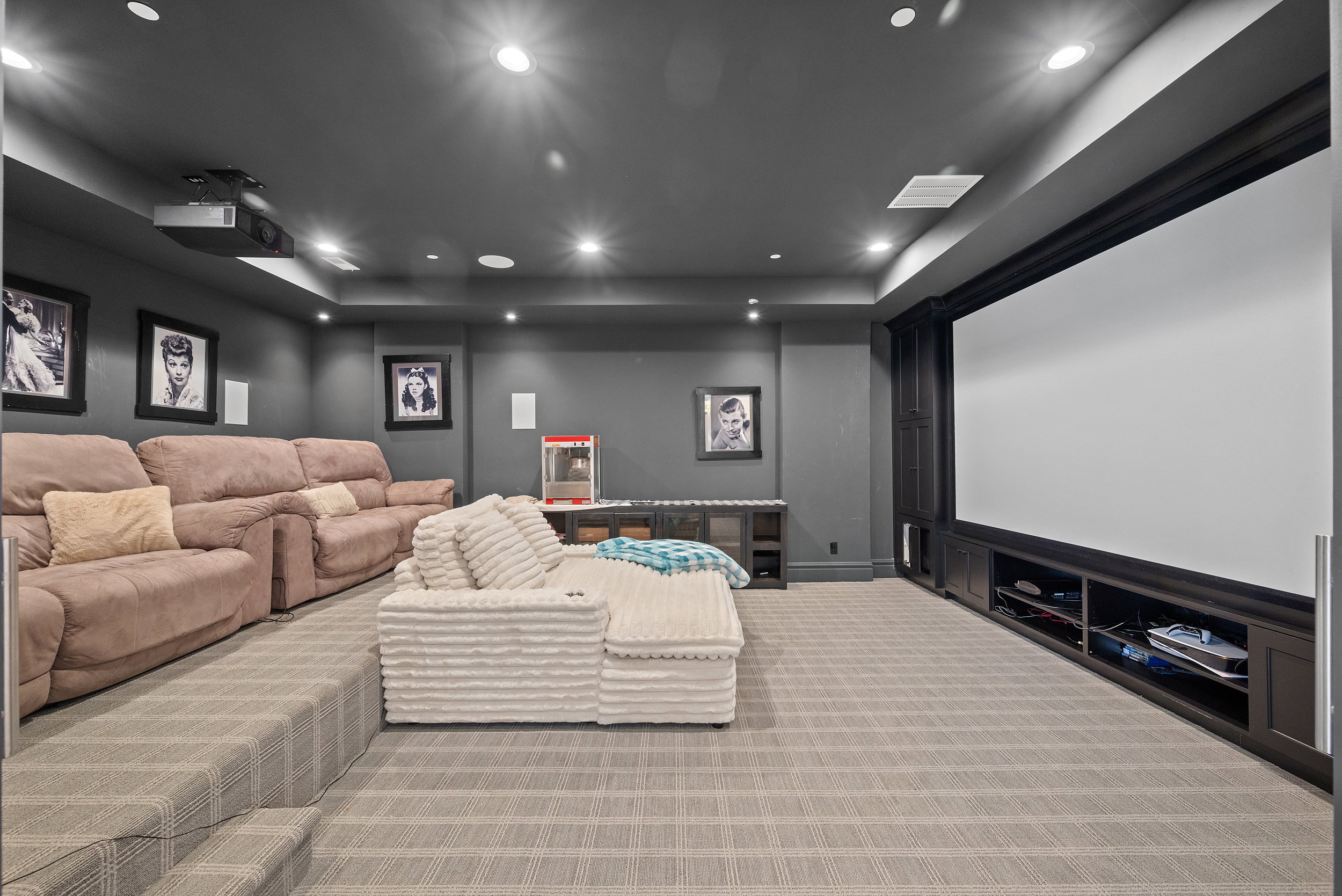 Dedicated media room featuring a large projection screen, tiered carpeted seating area, recessed ceiling lighting, and built-in dark wood cabinetry with open shelving