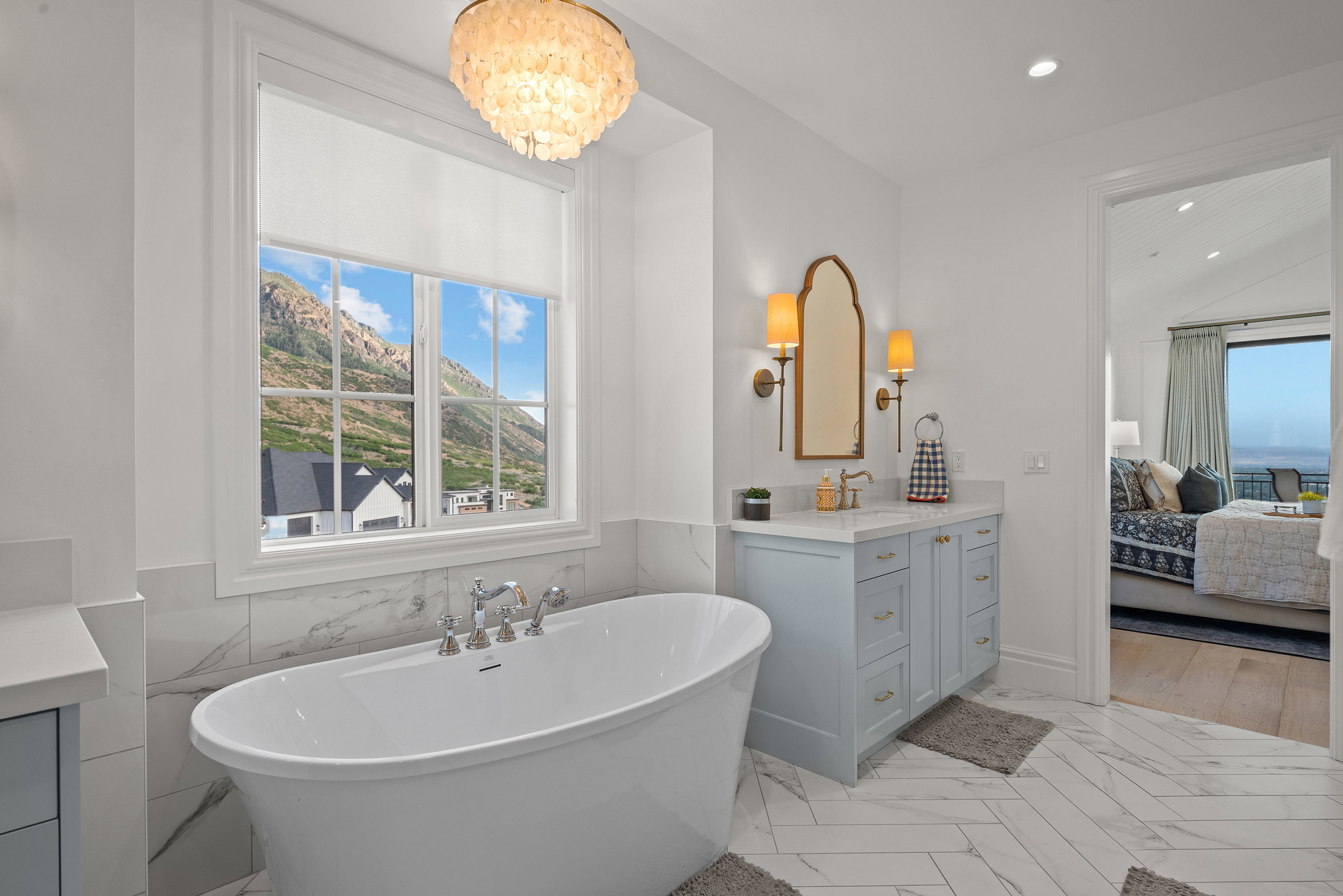 Bathroom featuring a freestanding soaking tub with deck-mounted fixtures, a window with mountain scenery, and a vanity with a light blue finish, a light-colored countertop, and a decorative mirror flanked by sconces