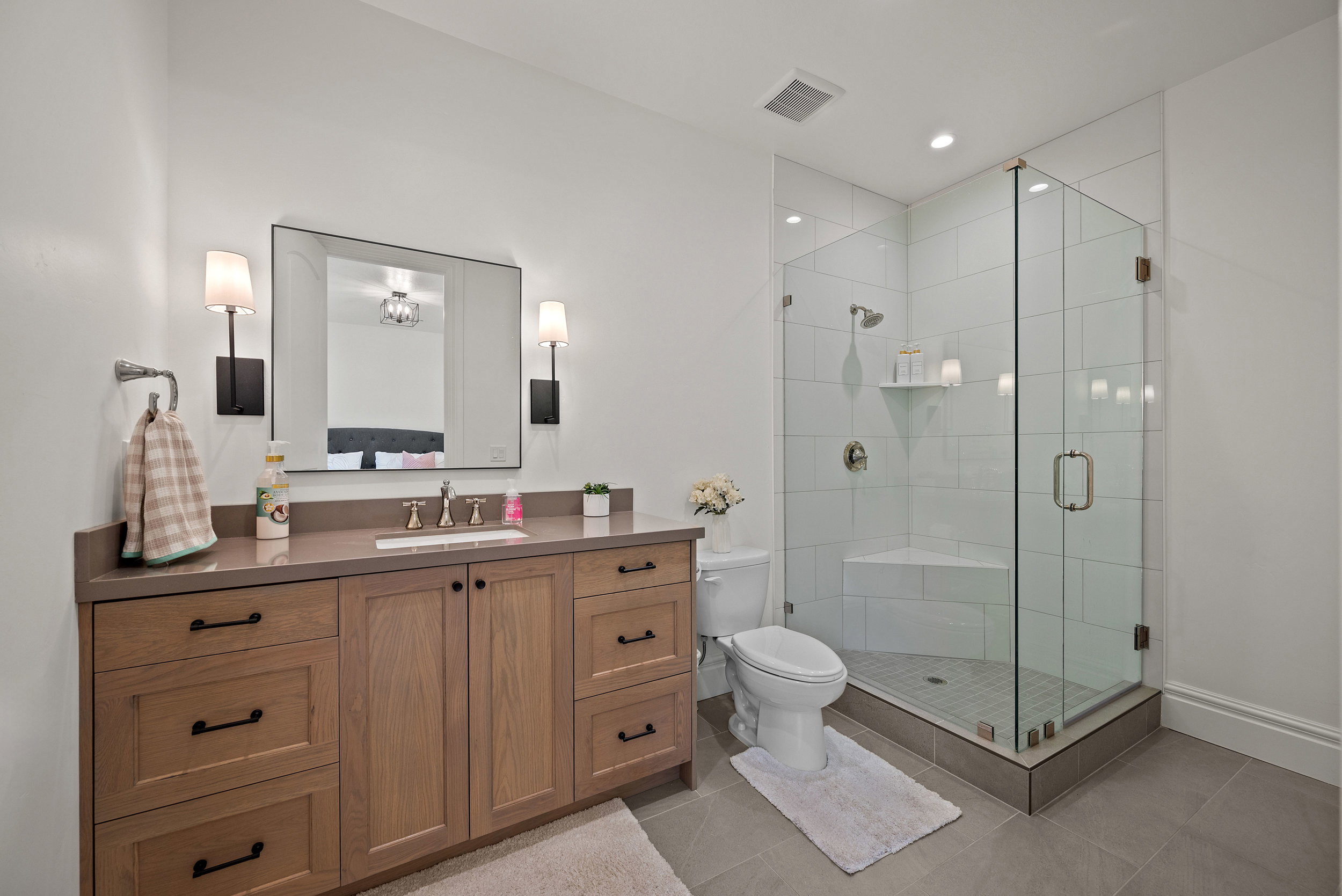 Contemporary bathroom featuring a glass-enclosed shower with a built-in bench, a single vanity with wood-finish cabinetry, a stone-finish countertop, and a large rectangular mirror with wall sconces