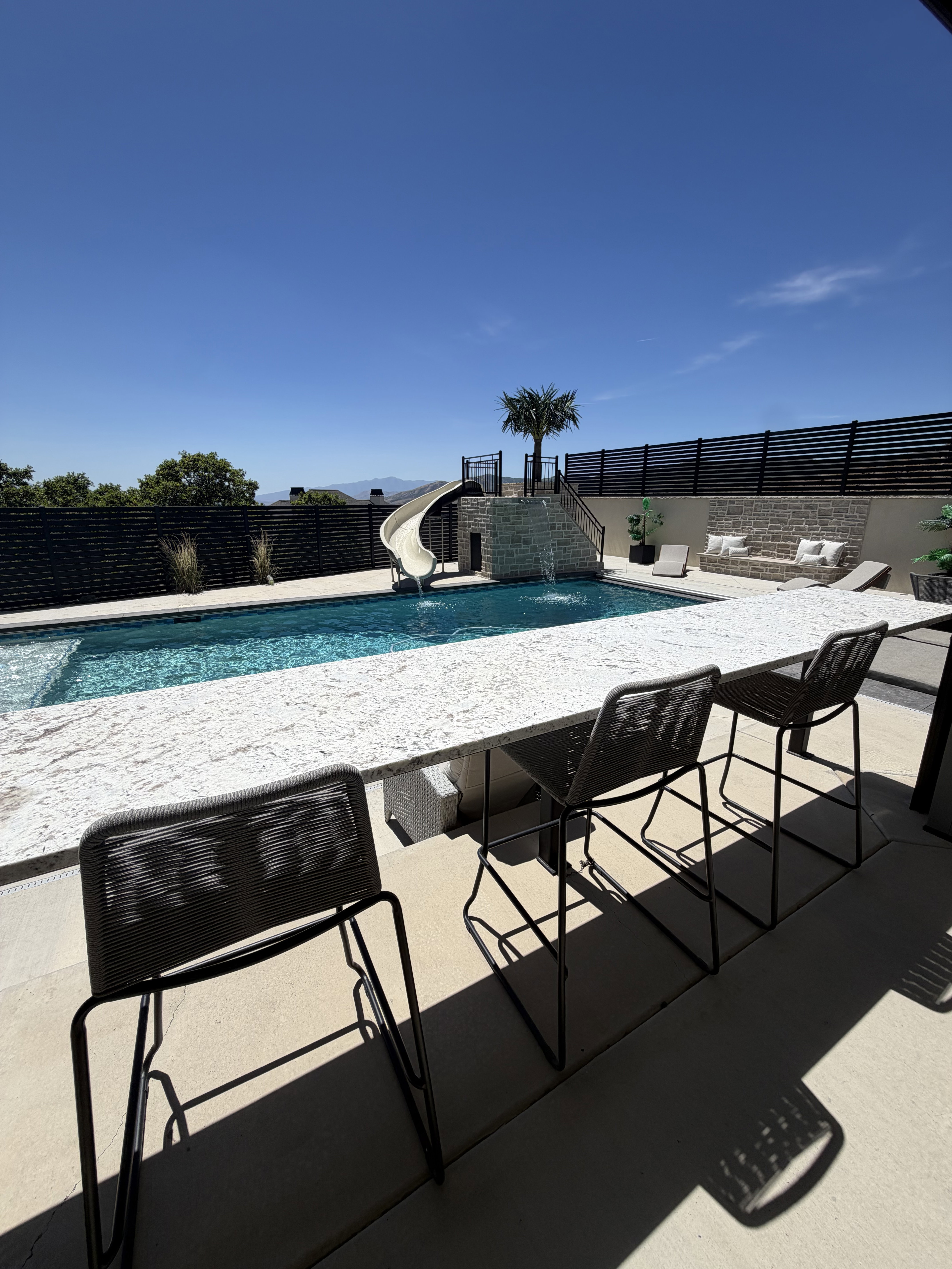Outdoor entertaining space featuring a built-in bar, pool with water feature and slide, stone retaining walls, and privacy fencing