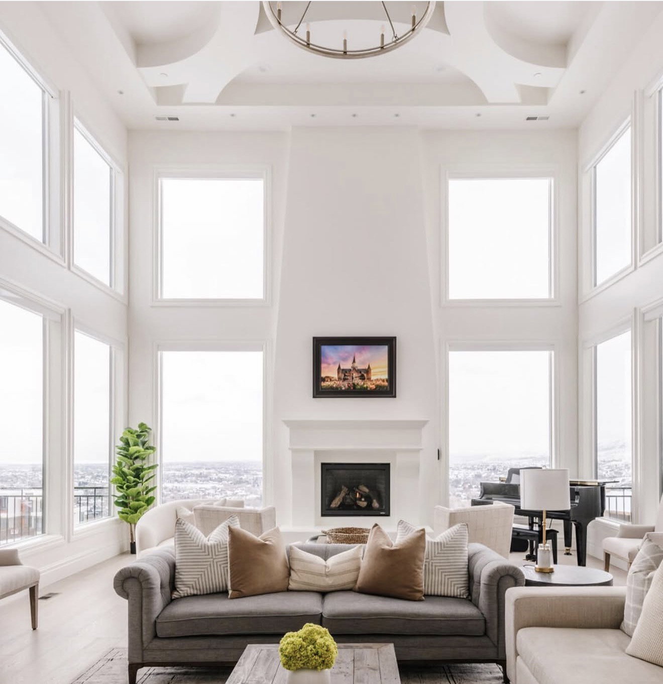 Spacious living area featuring a vaulted ceiling with a circular chandelier, a grand fireplace with a white mantel, and extensive windows showcasing distant scenery