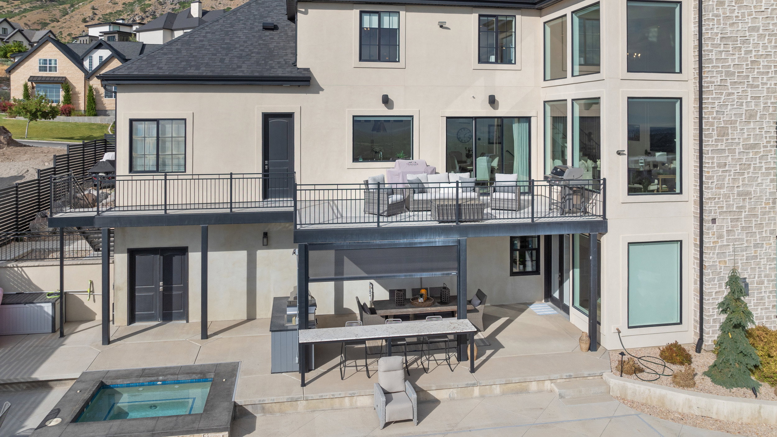 Multi-level stucco and stone exterior featuring a deck with black metal railing, lower patio with built-in outdoor kitchen, and an in-ground spa
