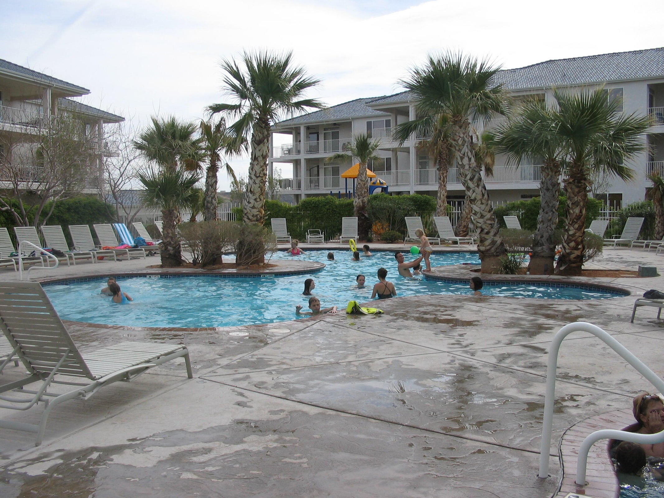 Expansive outdoor pool area featuring a structured concrete deck surround, integrated palm trees, and multiple chaise lounge chairs