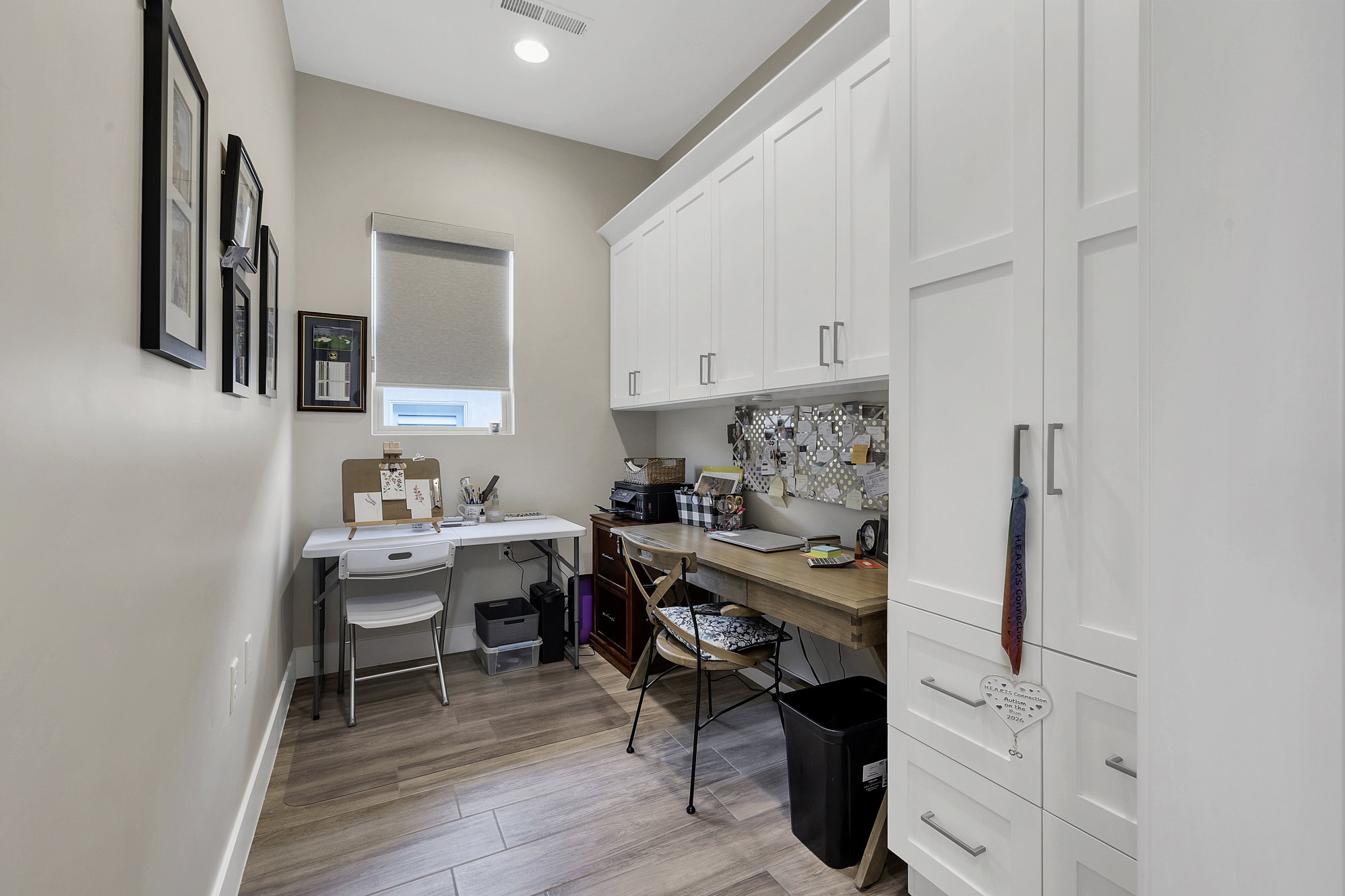 Dedicated workspace featuring extensive white cabinetry, a built-in wood-finish desk, wood-finish flooring, a window with a roller shade, and recessed lighting