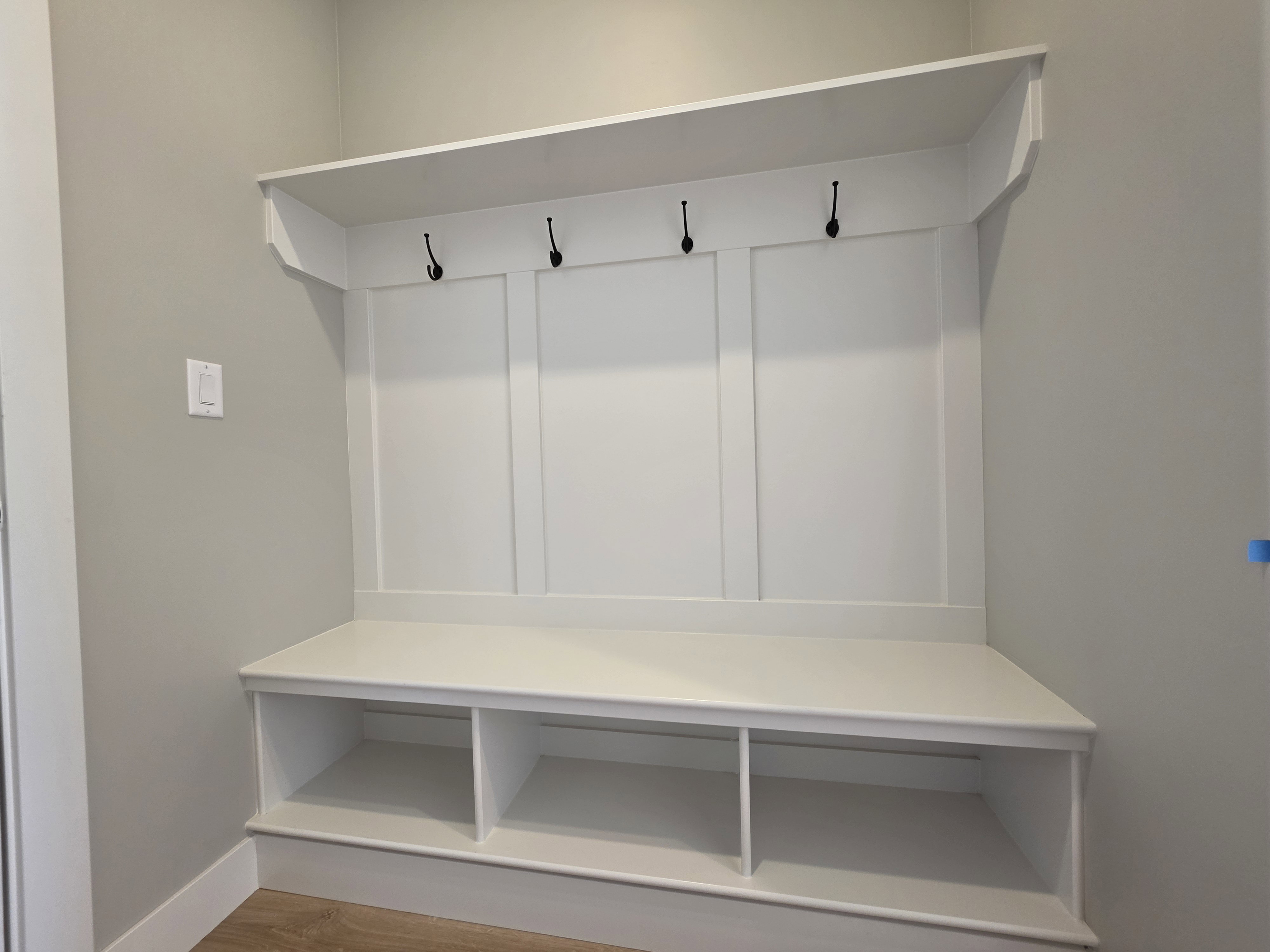 Built-in mudroom bench with three storage cubbies, white paneling, and four dark metal hooks