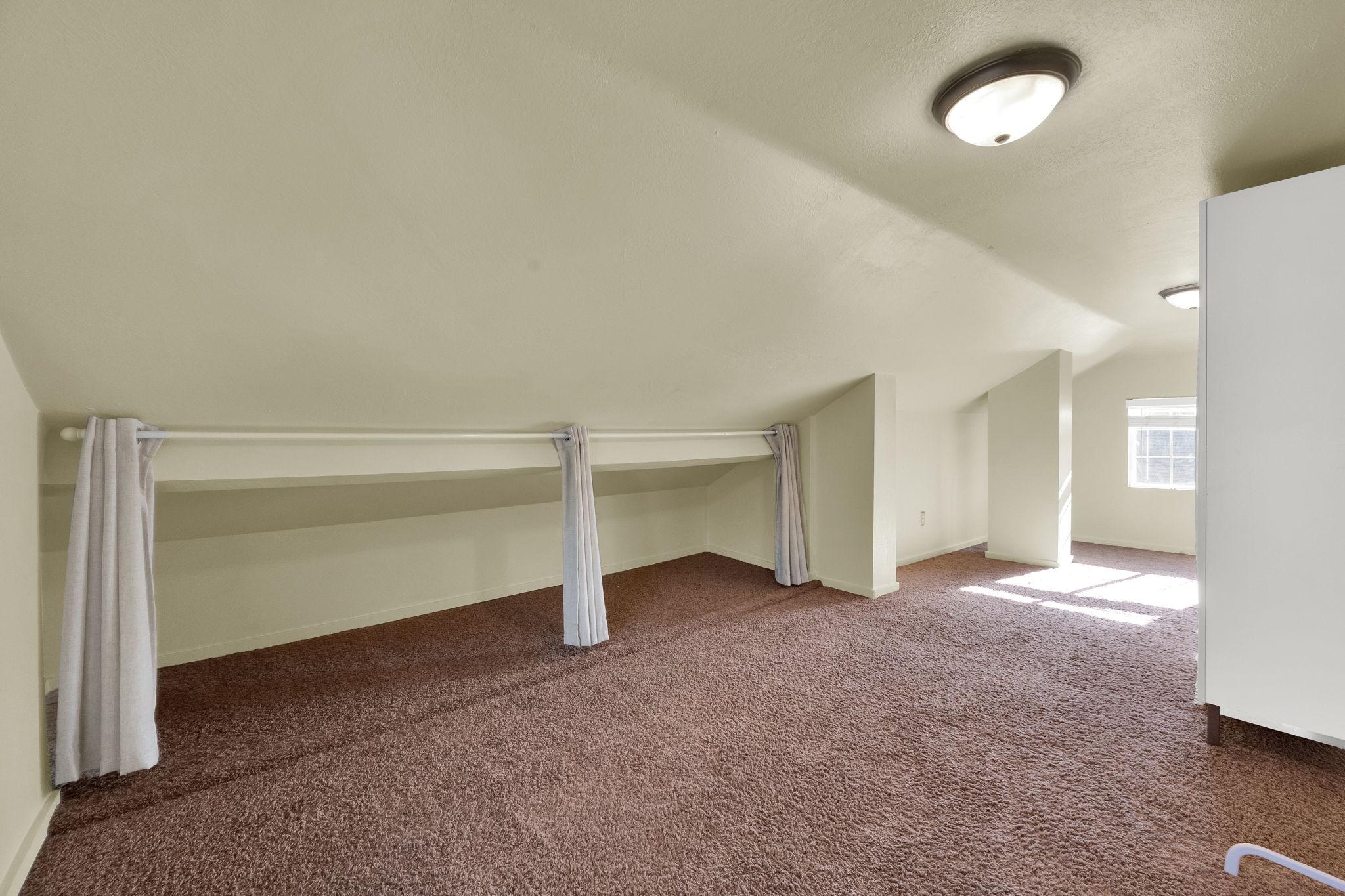 Carpeted attic space featuring sloped ceilings, recessed shelving with curtain accents, and natural light from a window