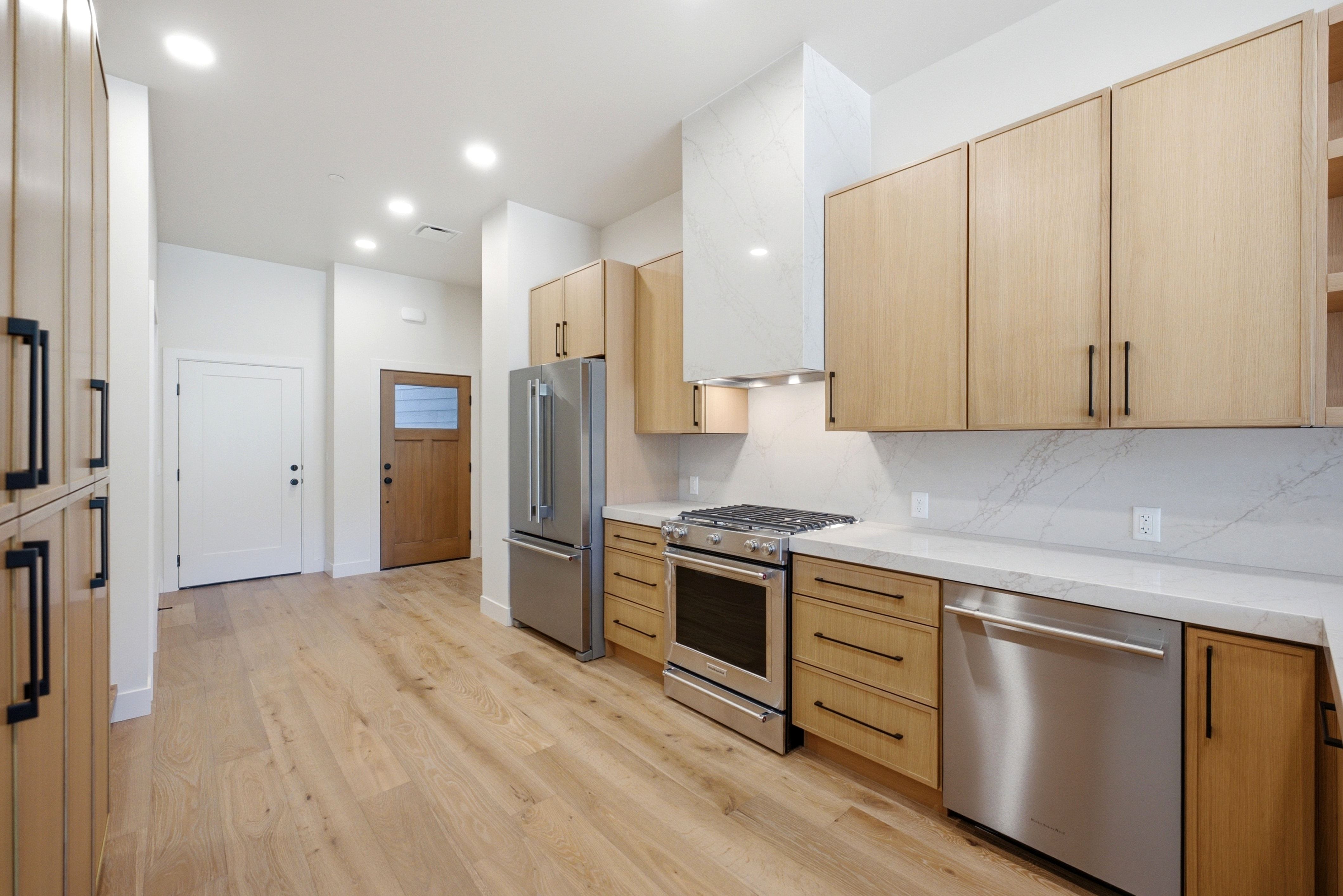 Modern kitchen featuring light wood-finish cabinetry with black hardware, stainless steel appliances, light-toned countertops, and a full-height backsplash