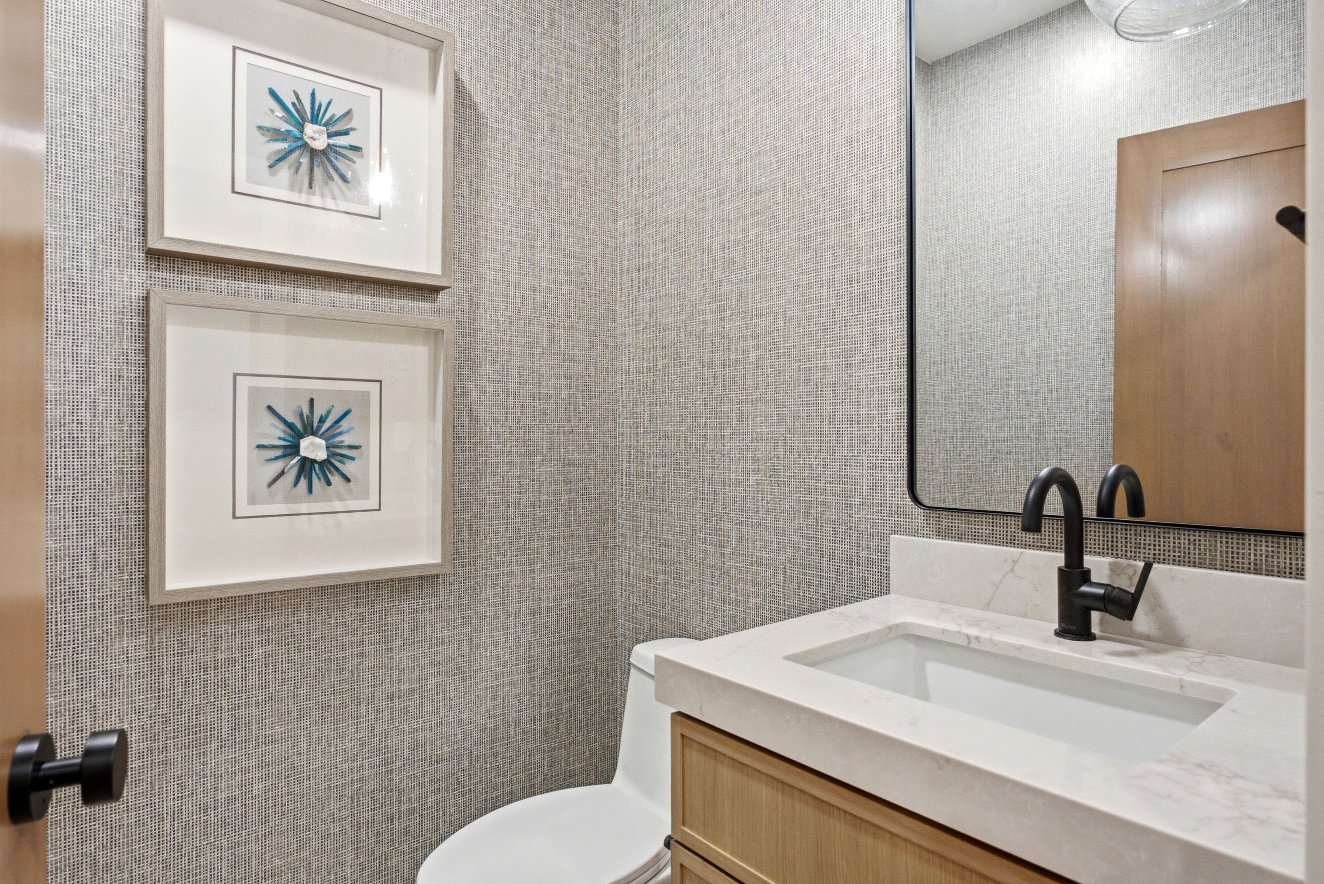 Powder room featuring textured wall covering, a light wood-finish vanity with an integrated rectangular sink, a light-toned countertop, and a matte black single-handle faucet