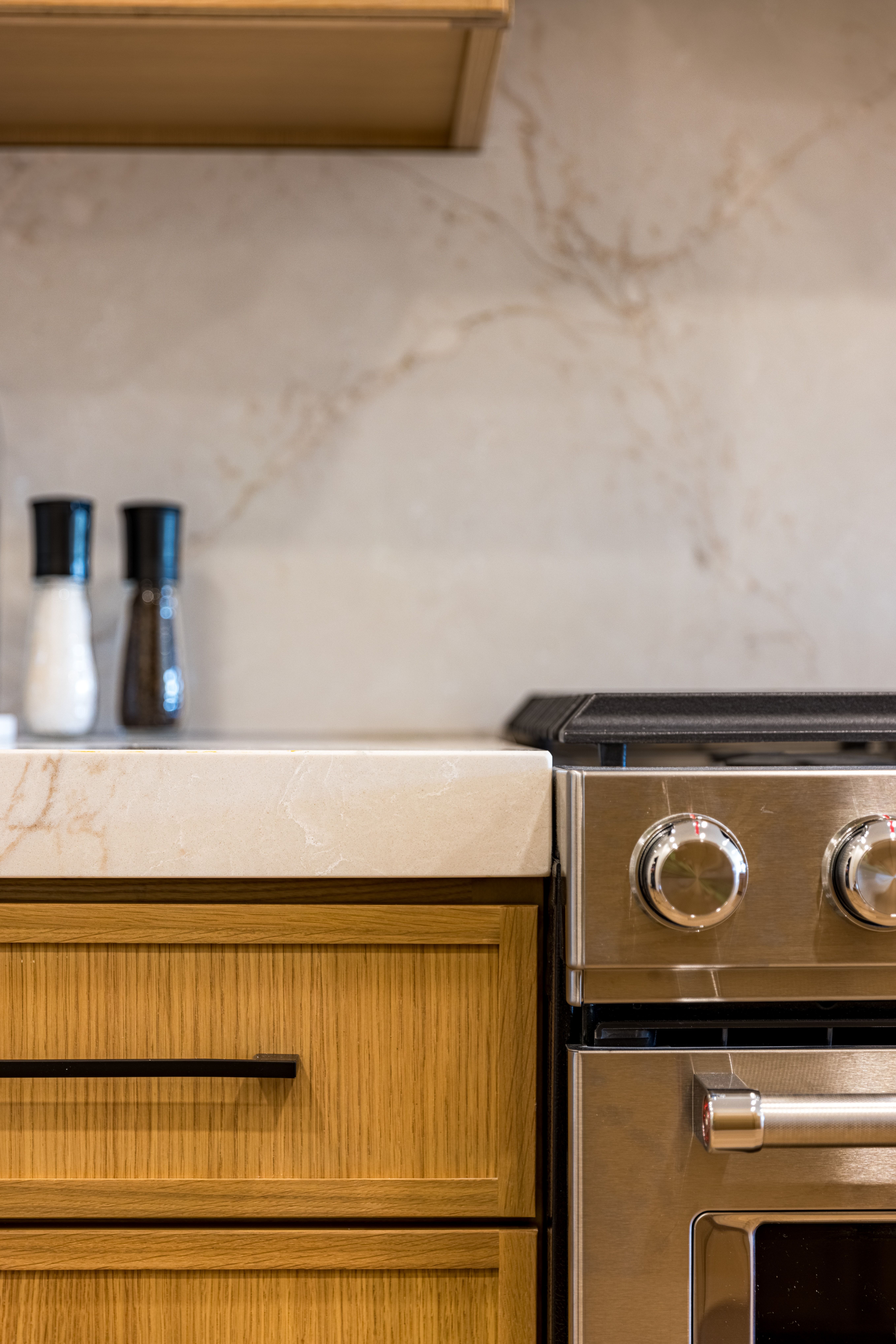 Stainless steel oven and cooktop with a light-toned countertop, wood cabinetry featuring horizontal grain and black hardware, and a marbled backsplash