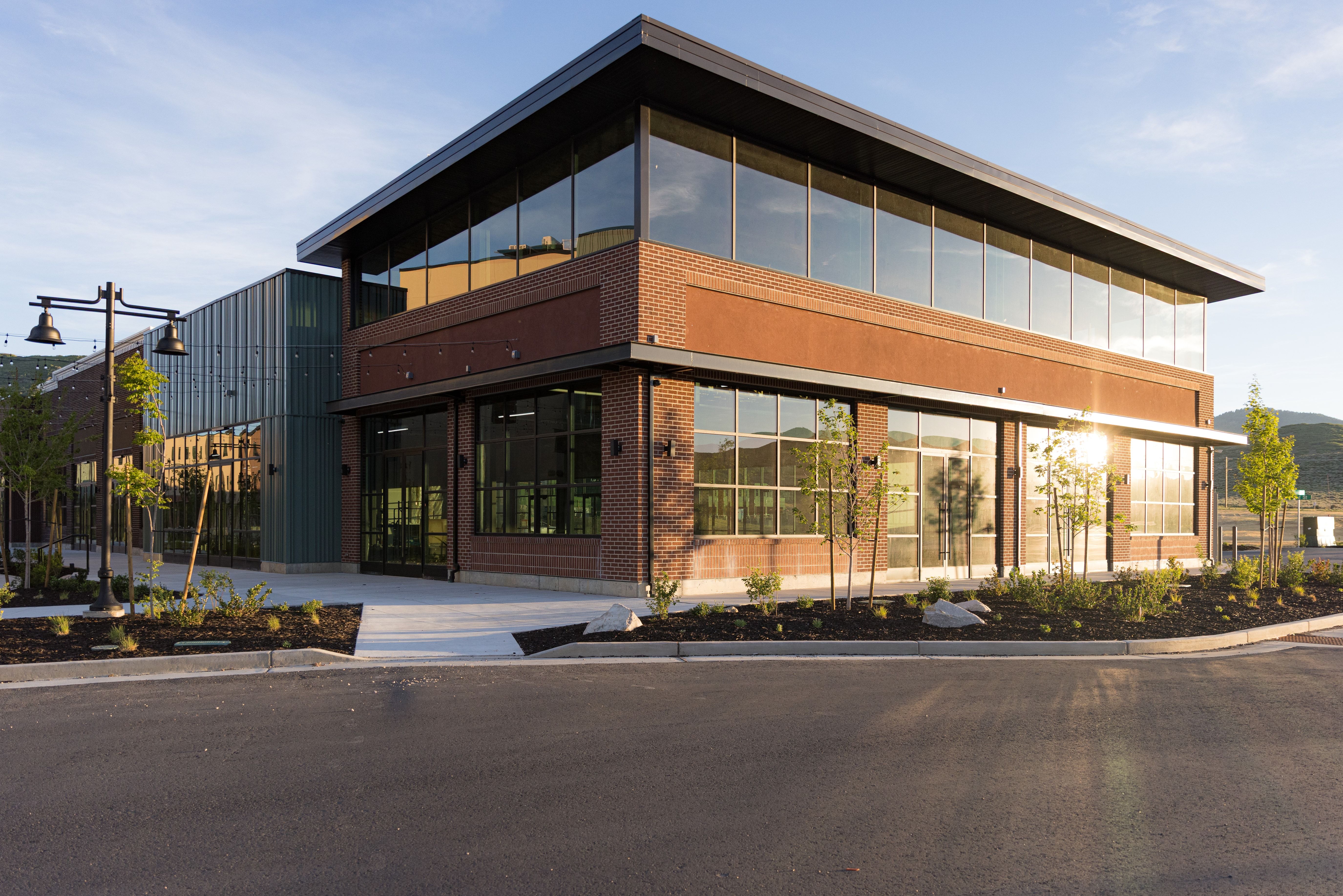 Modern commercial building featuring brick and metal panel exteriors, extensive window lines, a prominent cantilevered roofline, and landscaped grounds with young trees and shrubs