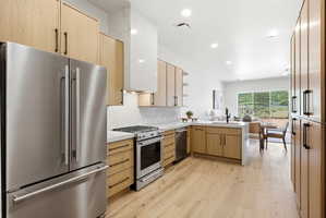 Open-concept kitchen featuring light wood-finish cabinetry, stainless steel appliances, a range hood with a white stone accent, and a light wood-finish flooring