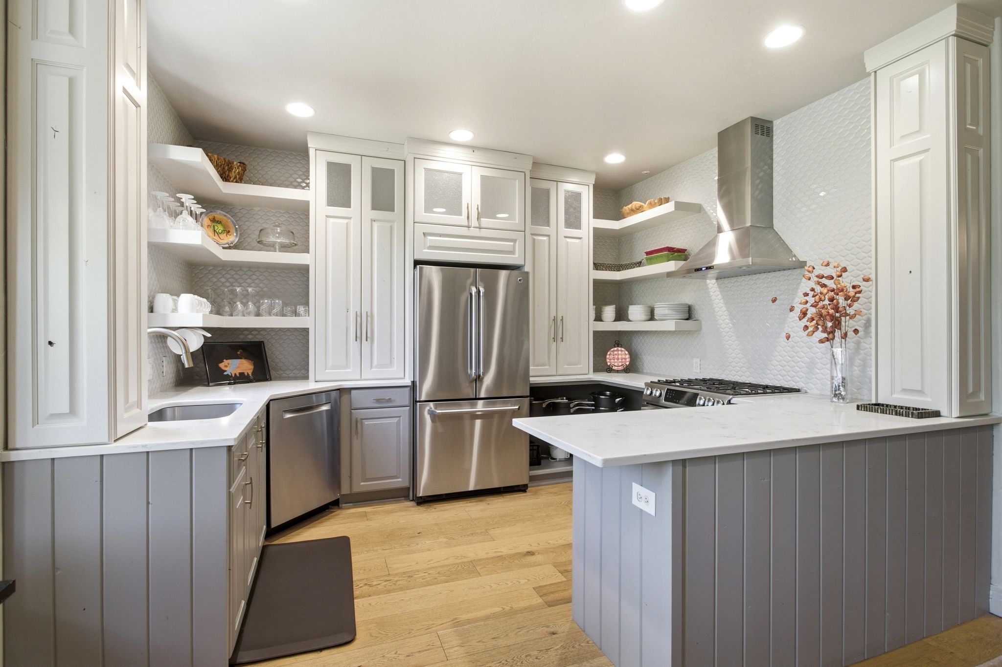 Kitchen featuring light wood-finish flooring, white perimeter cabinetry with glass-front accents, and a stainless steel appliance suite including a refrigerator, dishwasher, and range with a hood vent