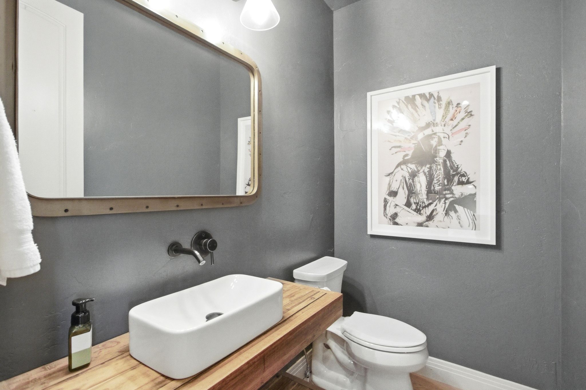 Contemporary bathroom featuring a vessel sink, wall-mounted faucet, wood countertop, industrial-style mirror, and a white toilet