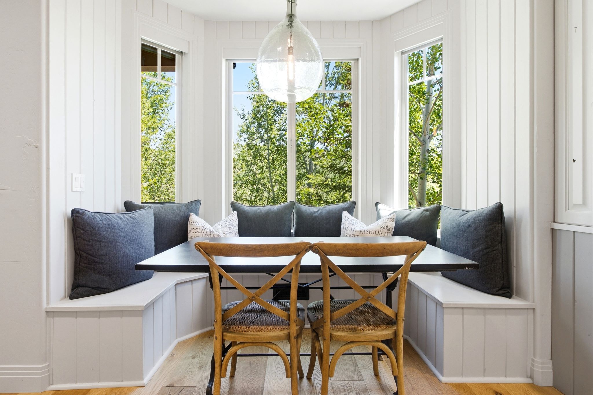 Built-in corner bench seating with white paneling, multiple large windows, a black table with a dark metal base, an overhead glass pendant light fixture, and wood-finish flooring