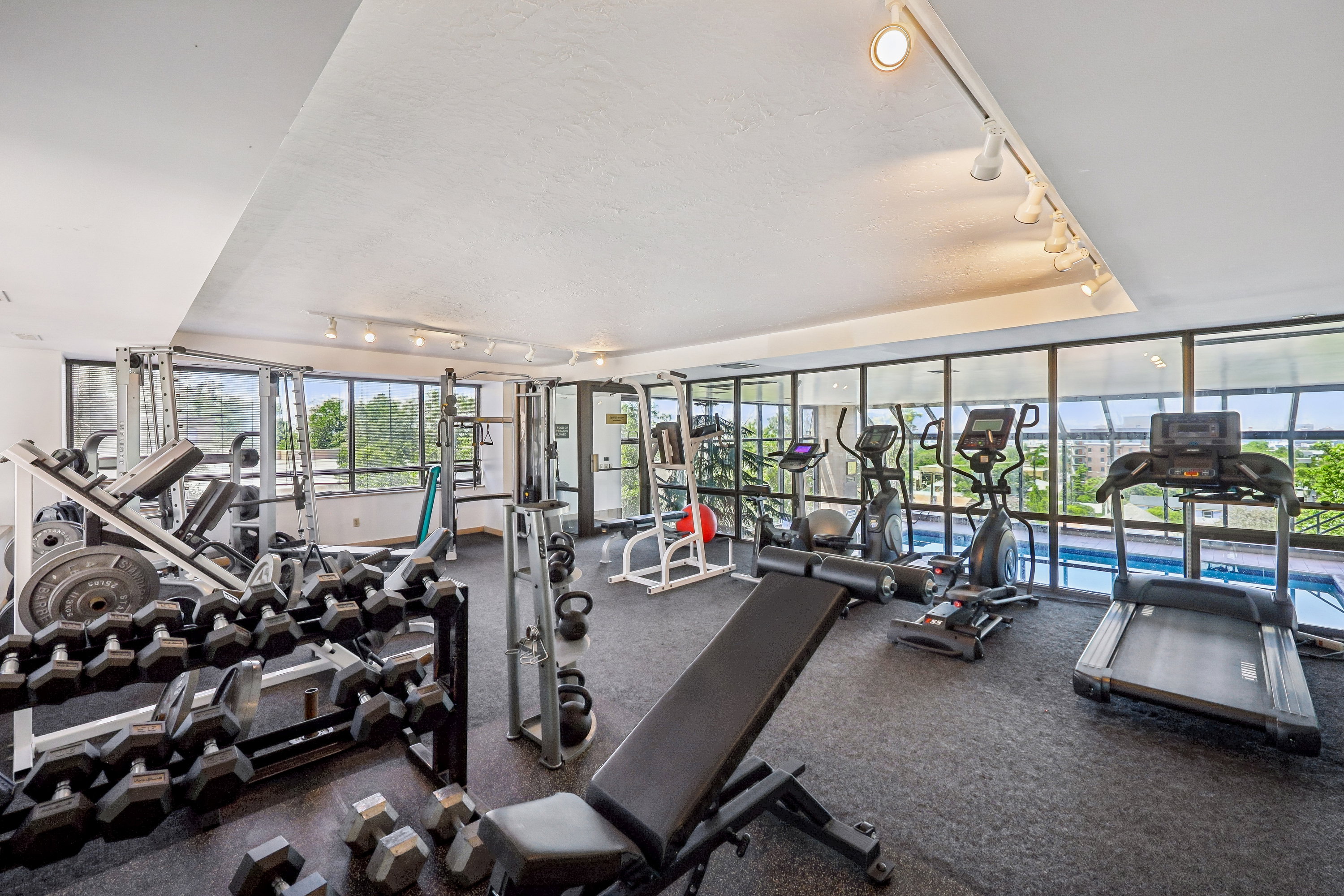 Fitness center with track lighting, expansive windows, cushioned flooring, and an adjacent indoor pool