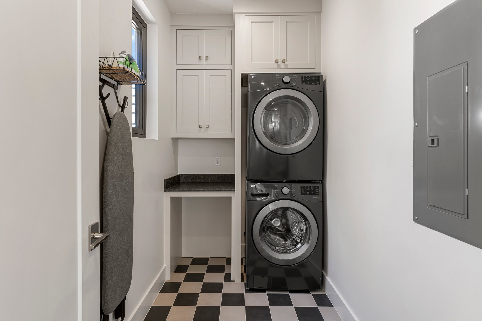 Laundry area featuring stacked appliances, black and white checkered flooring, a built-in counter with dark stone-finish surface, and white upper cabinetry