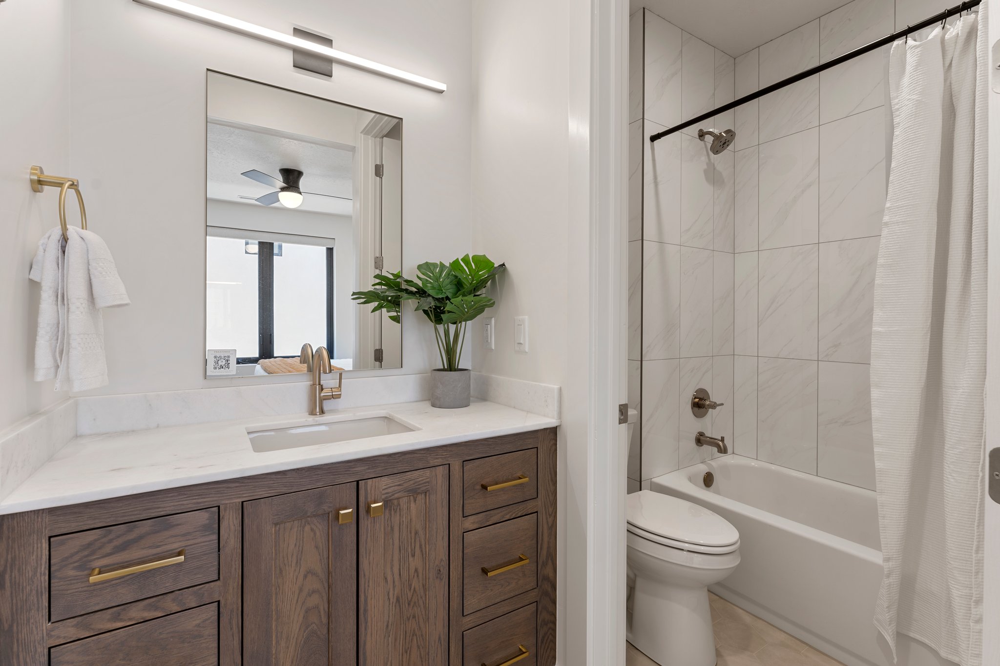 Bathroom featuring a wood-finish vanity with gold-tone hardware, a white countertop with an integrated rectangular sink, a wall-mounted rectangular mirror, and a linear overhead light fixture