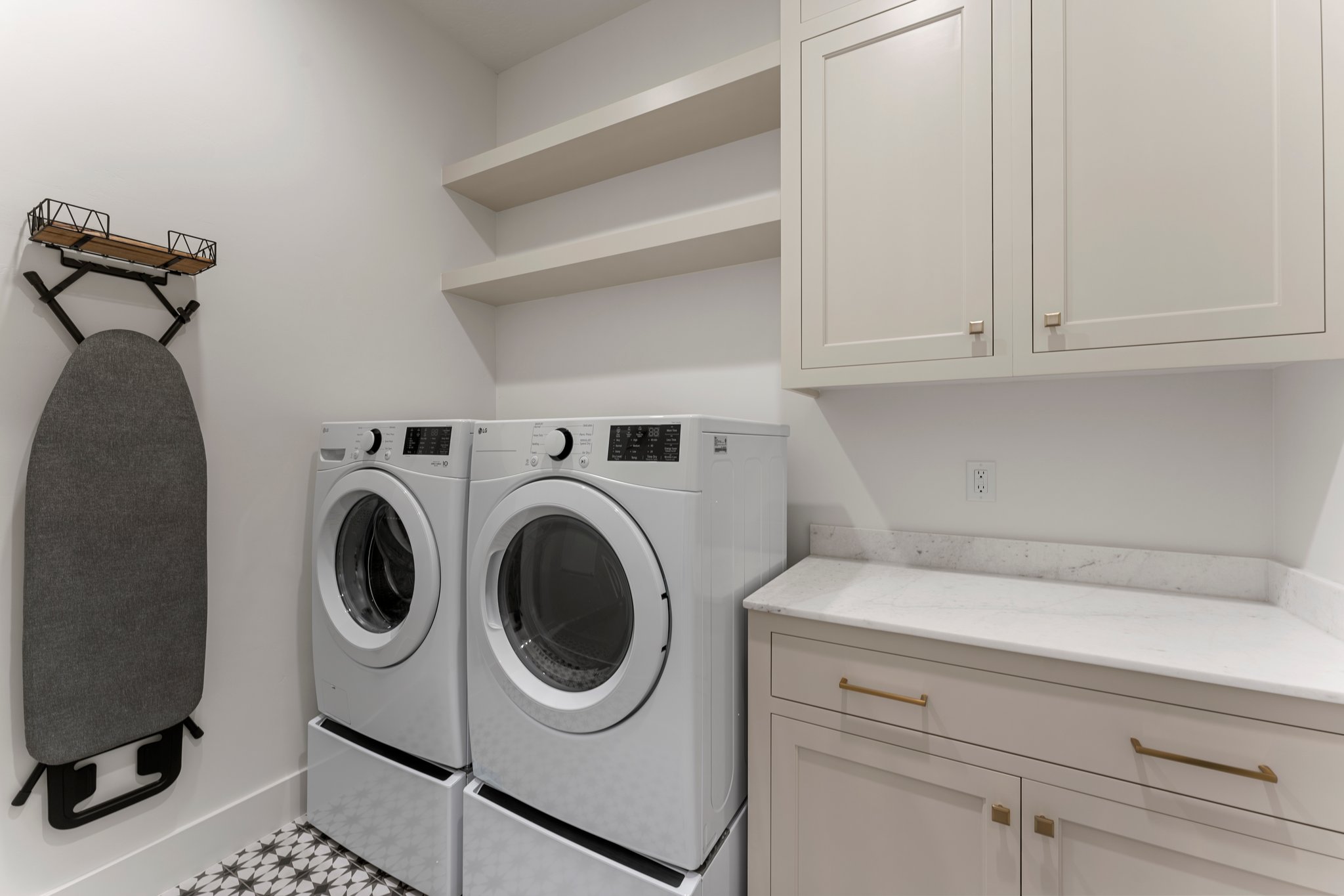 Laundry area featuring patterned floor tiles, light-toned shaker-style cabinetry with gold-tone hardware, white countertop, and open shelving