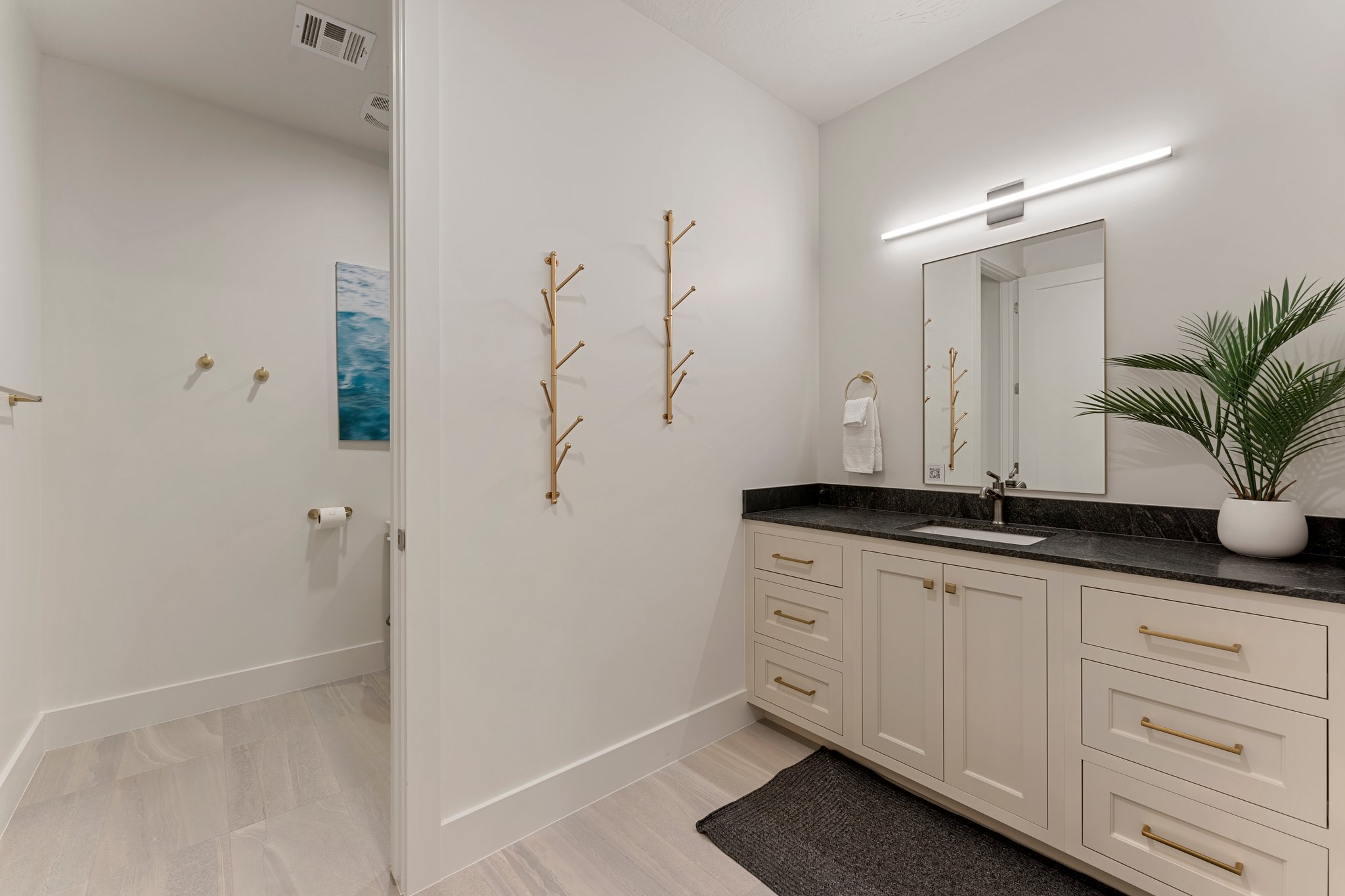 Bathroom featuring a light wood-finish floor, a long vanity with cream cabinetry, gold hardware, a dark countertop, and an undermount sink with a wall-mounted mirror