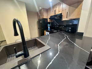 Kitchen featuring dark stone countertops with prominent white veining, a deep single-basin sink with a matte black high-arc faucet, light wood-finish cabinetry with dark hardware, a black electric range with an overhead vent, and a black refrigerator