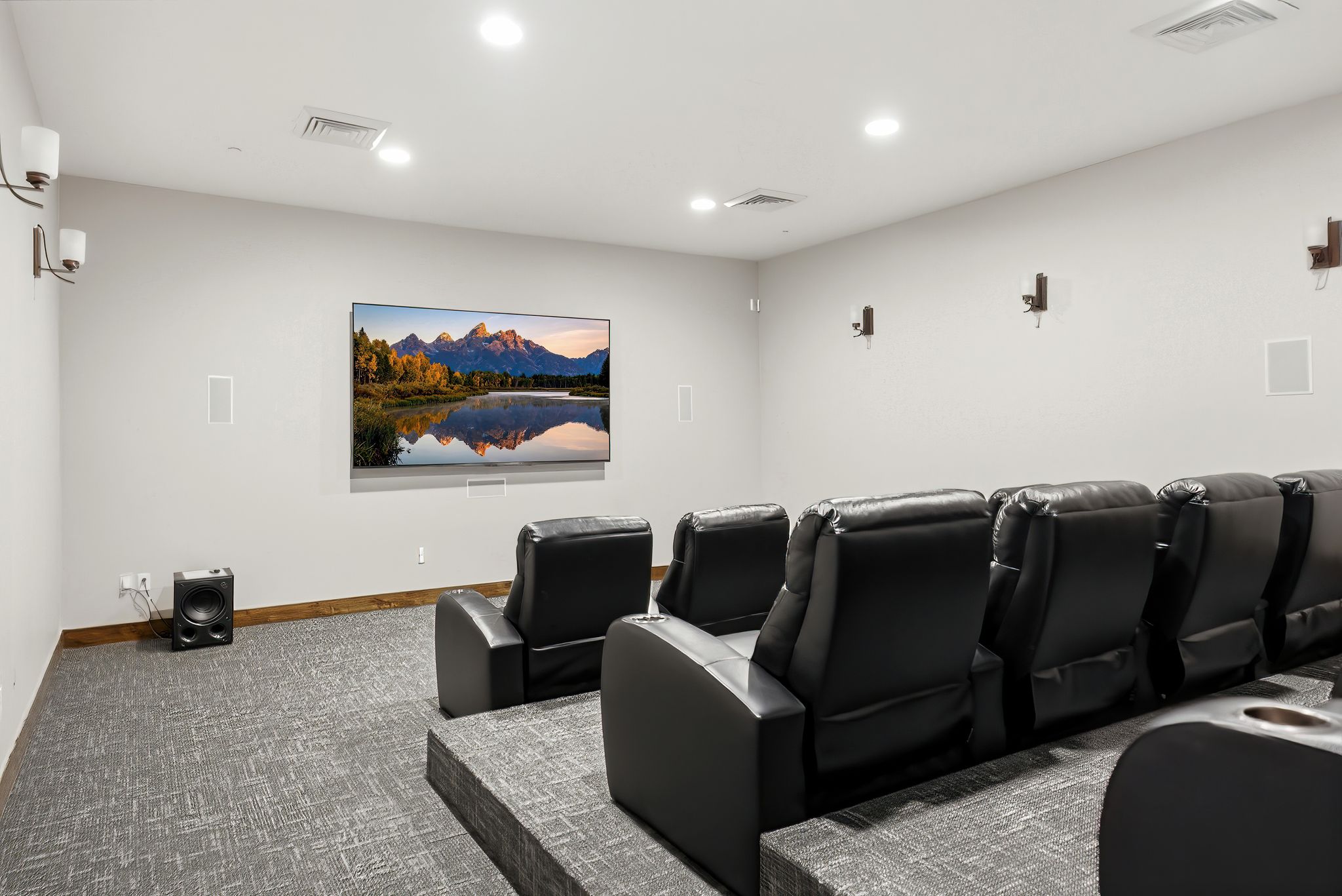 Dedicated media room featuring a large wall-mounted screen, tiered seating platforms, wall-mounted light fixtures, recessed ceiling lighting, and wall-integrated speakers