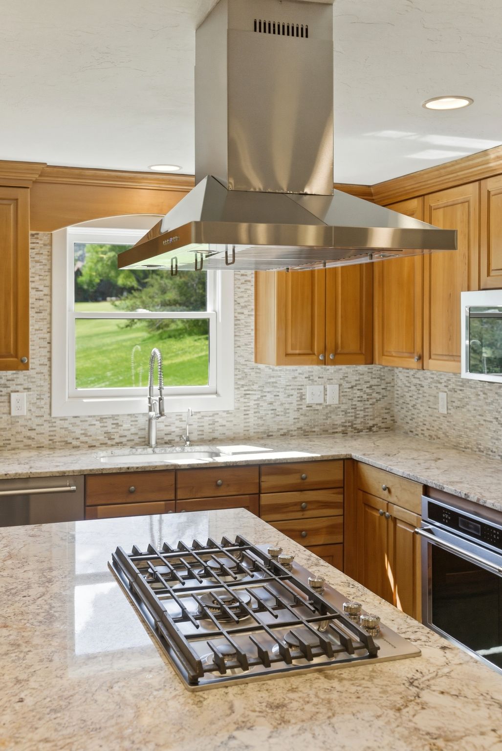 Kitchen featuring a stainless steel range hood, light wood cabinetry, stone countertops, a tile backsplash, and a built-in cooktop