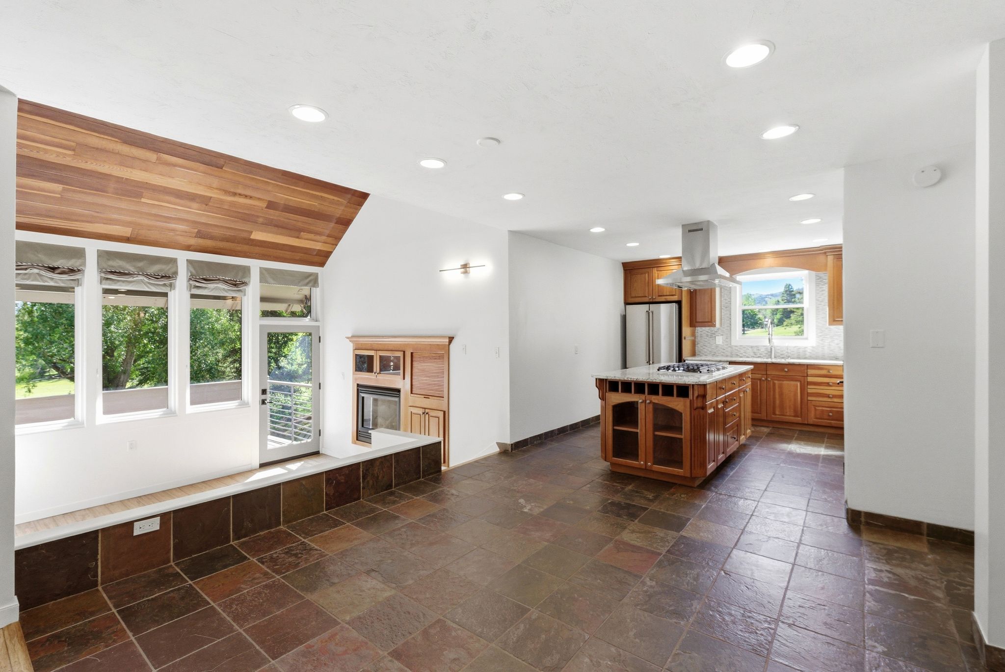 Spacious kitchen and living area featuring natural wood cabinetry, a central island with integrated cooktop, stainless steel appliances, and a tiled backsplash