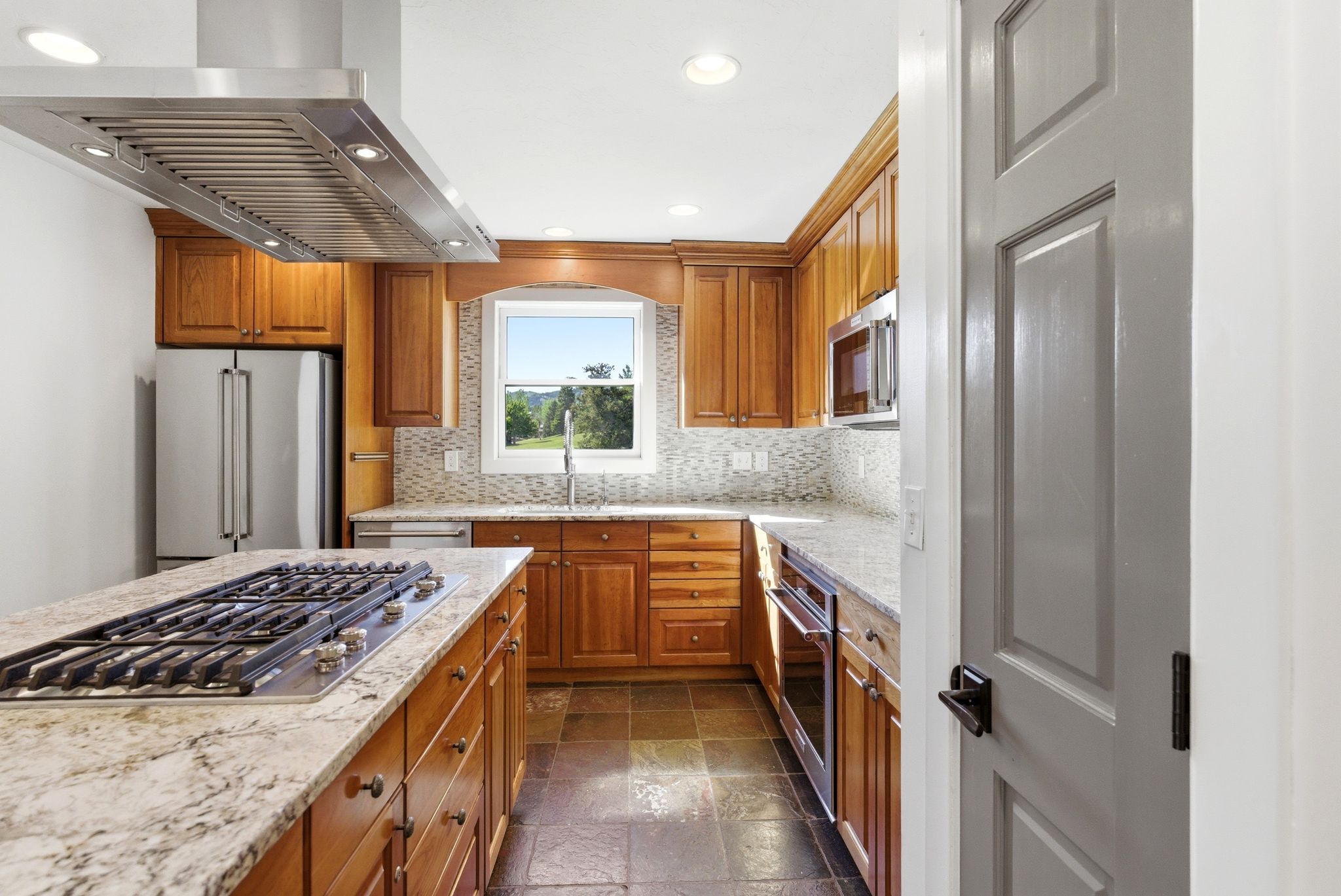 Kitchen featuring stainless steel appliances, wood cabinetry, stone countertops, a tile backsplash, and slate flooring