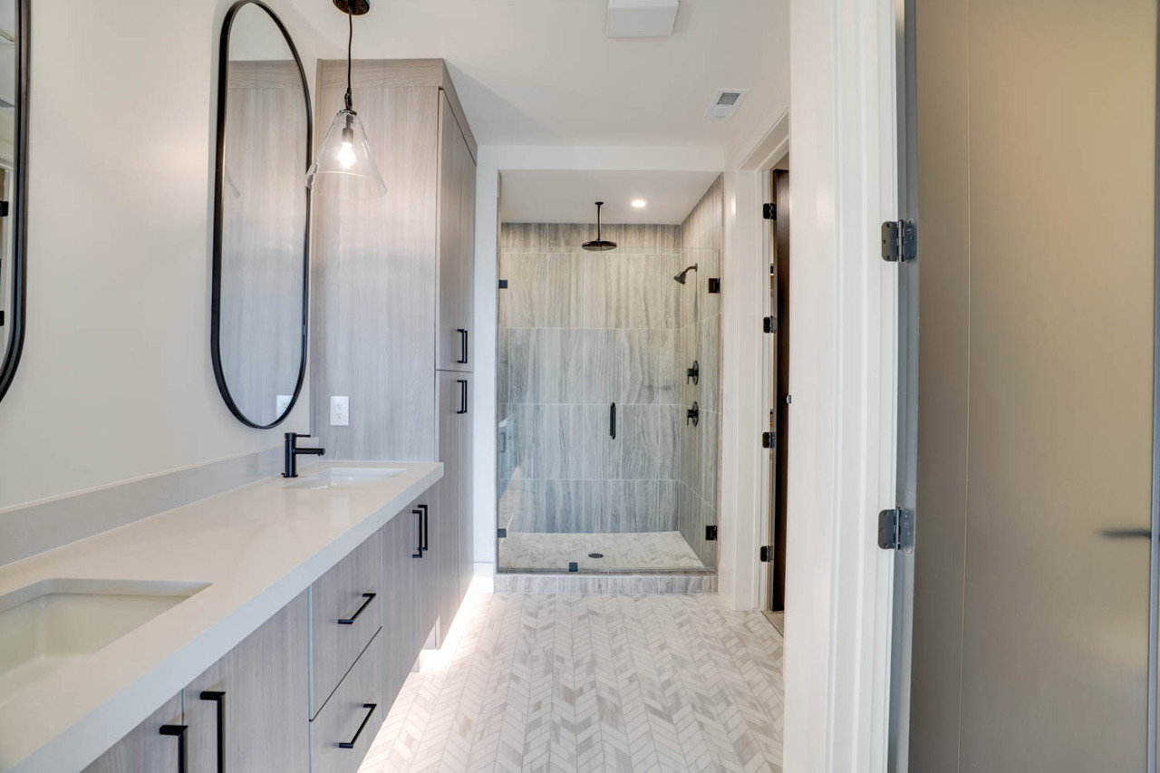 Dual vanity with integrated sinks and matte black fixtures, full-height custom cabinetry, glass-enclosed shower with multiple showerheads, and herringbone-patterned tile flooring