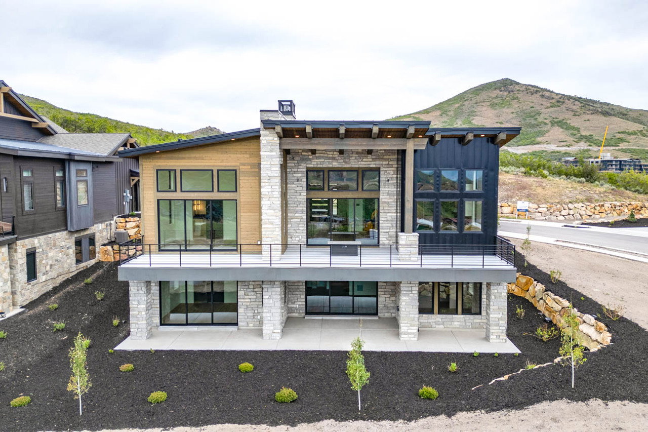 Modern multi-level residence featuring a stone and wood-panel exterior, extensive full-height windows, a prominent upper-level balcony with metal railings, and a lower-level patio