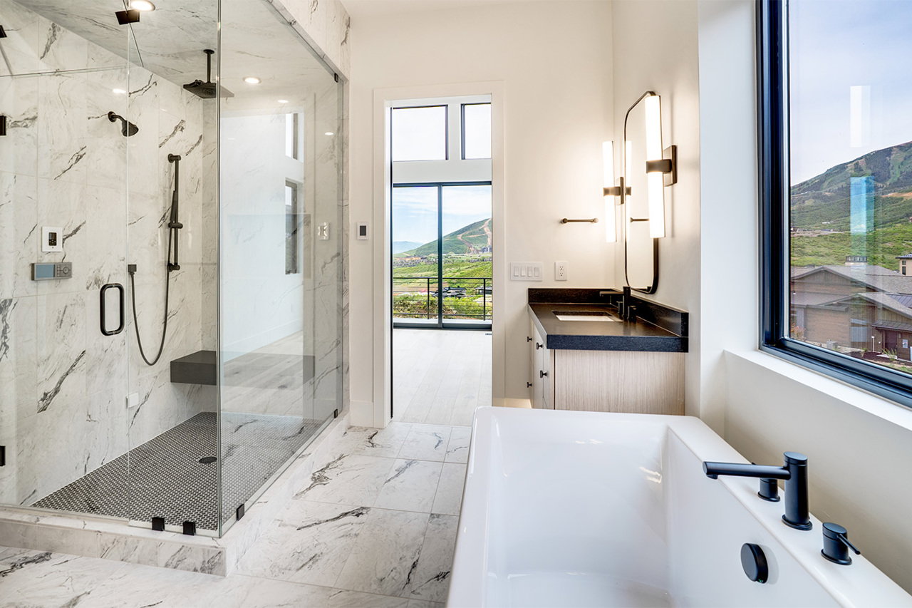 Spacious bathroom featuring a freestanding tub with matte black fixtures, a walk-in shower with a built-in bench, marble-finish wall and floor tile, a modern vanity with a dark countertop, and a large window overlooking a mountain landscape