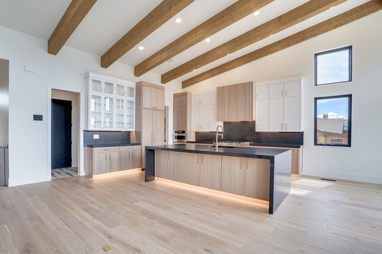 Modern kitchen featuring exposed wood beams, light wood-finish flooring, a waterfall island with integrated lighting, a matte black countertop and backsplash, and a combination of light wood and white cabinetry